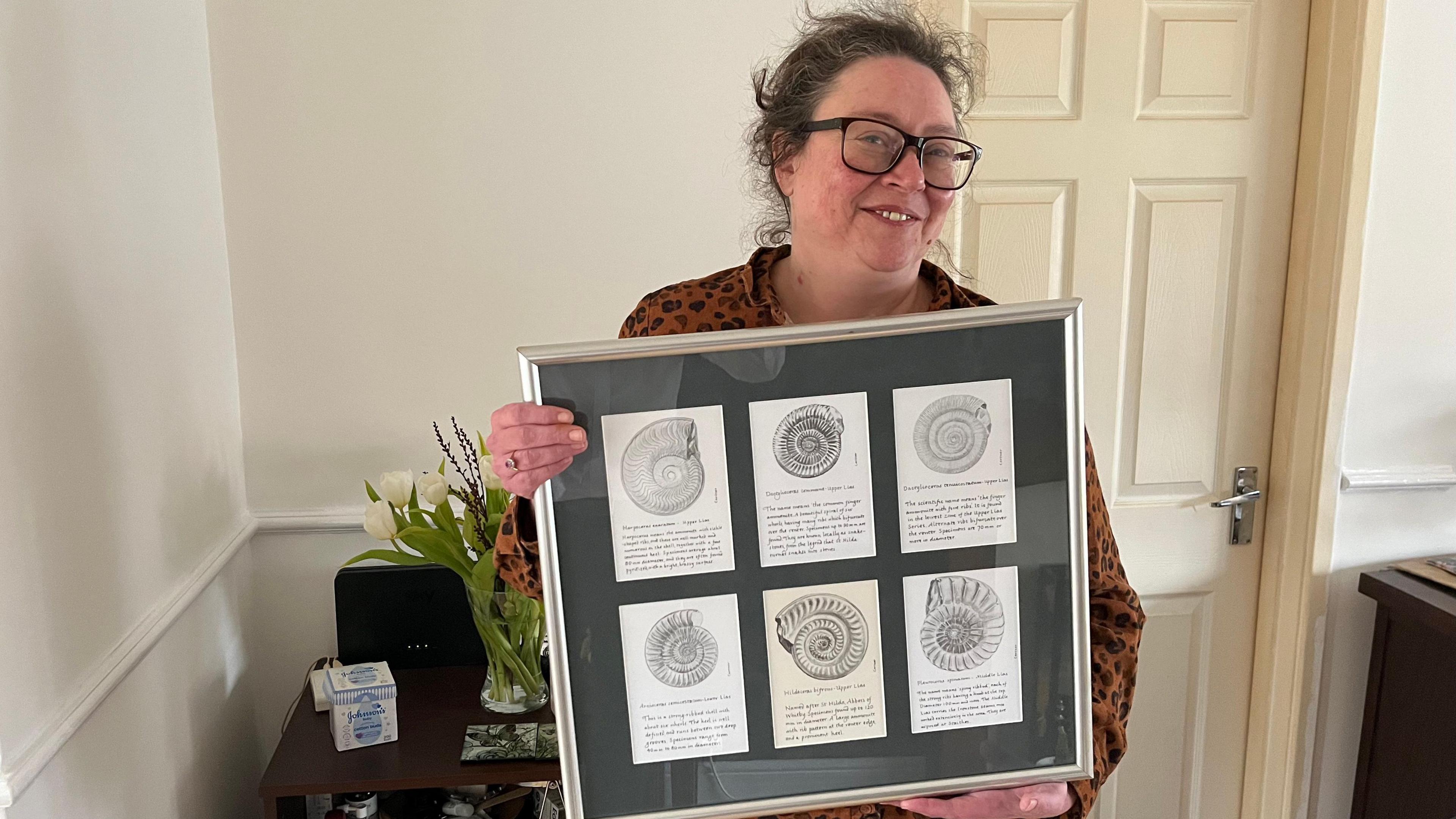 Helen Clay. She is standing in her home holding a framed collection of her grandfather's ammonite sketches. She is smiling at the camera and wearing glasses.