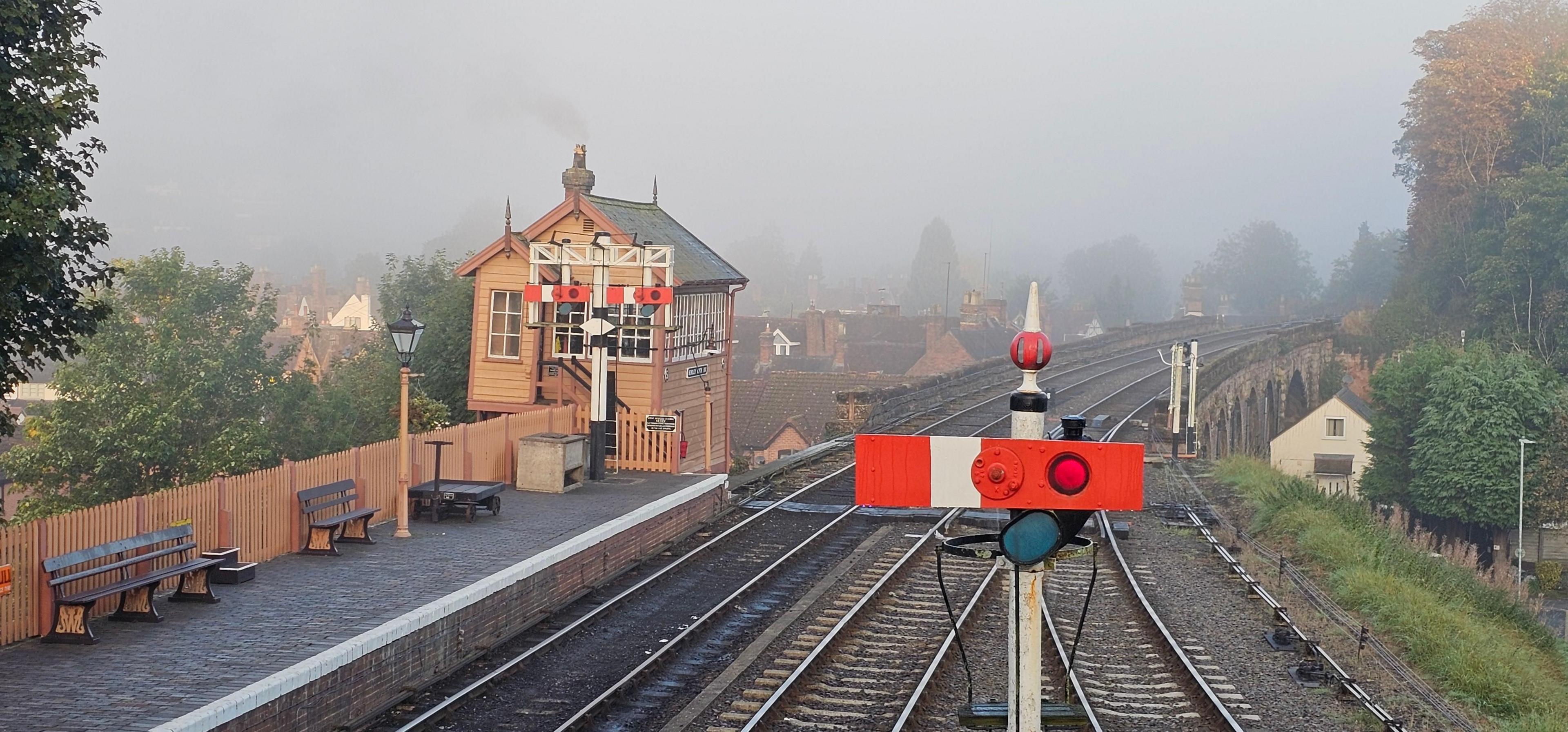 A railway signal is in the foreground, with a platform with benches further back and behind that a historic signal box. The rails disappear into the distance over a bridge. 