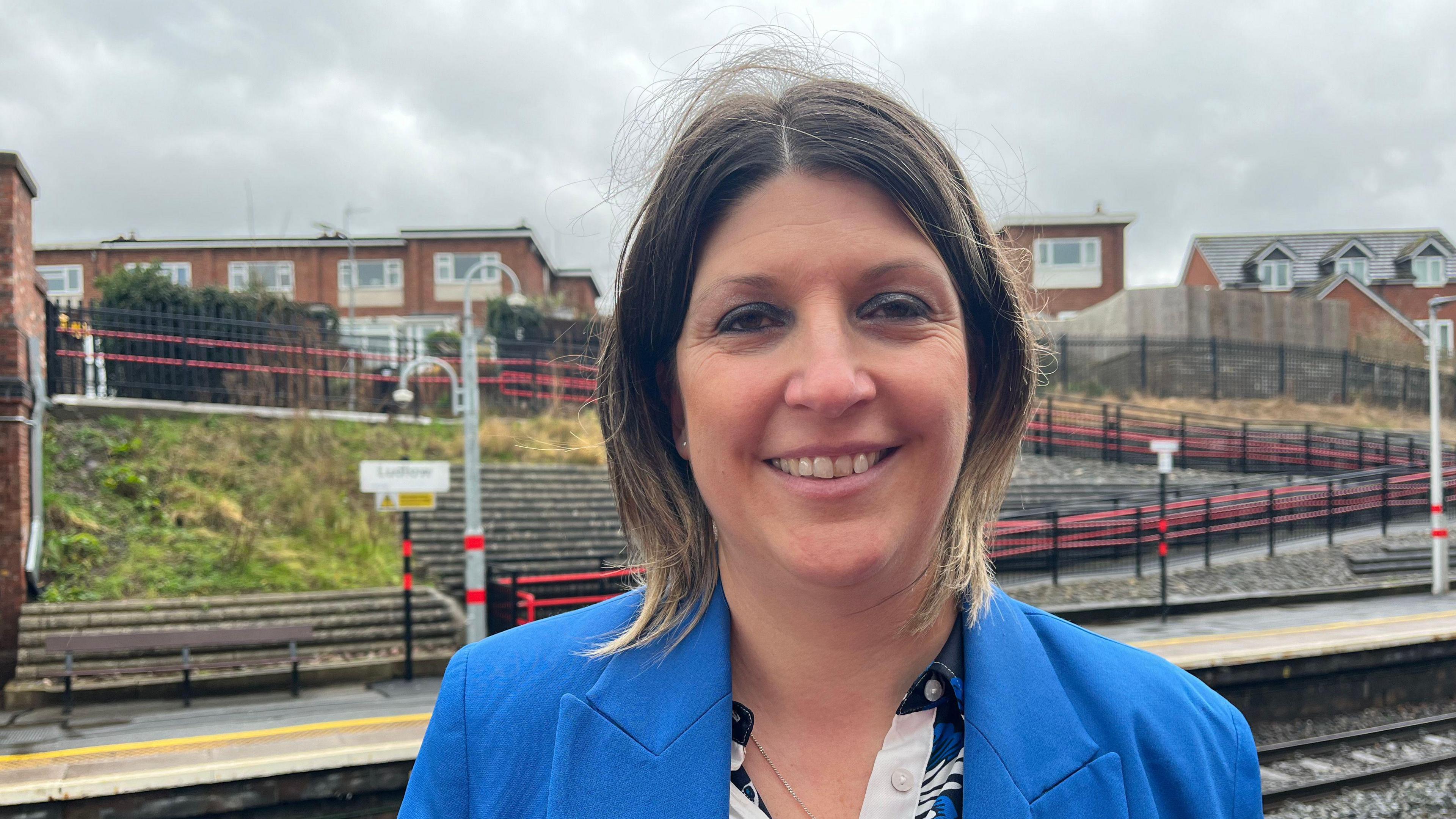 Rachel pictured smiling and looking into the camera. She's wearing a bright blue blazer and a white, black, and blue patterned shirt. She's stood on one of the platforms at Ludlow station, with houses visible just beyond the station boundaries. The sky is grey and overcast.