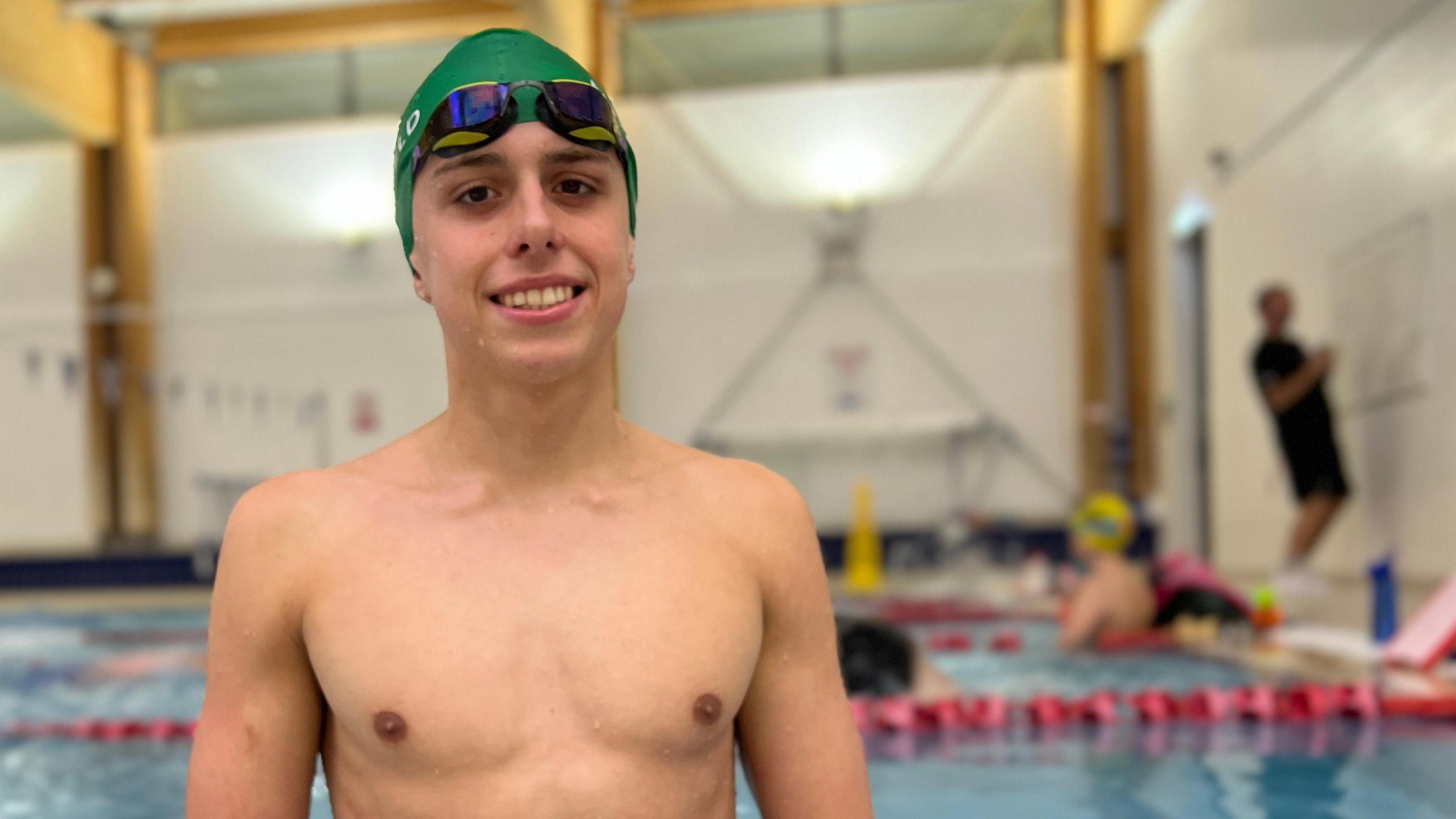 Filip smiles at the camera as he stands in a swimming pool. He has a green swimming cap and goggles on his head. Behind him other swimmers are resting at the end of the lane while a coach writes instructions on a white board.