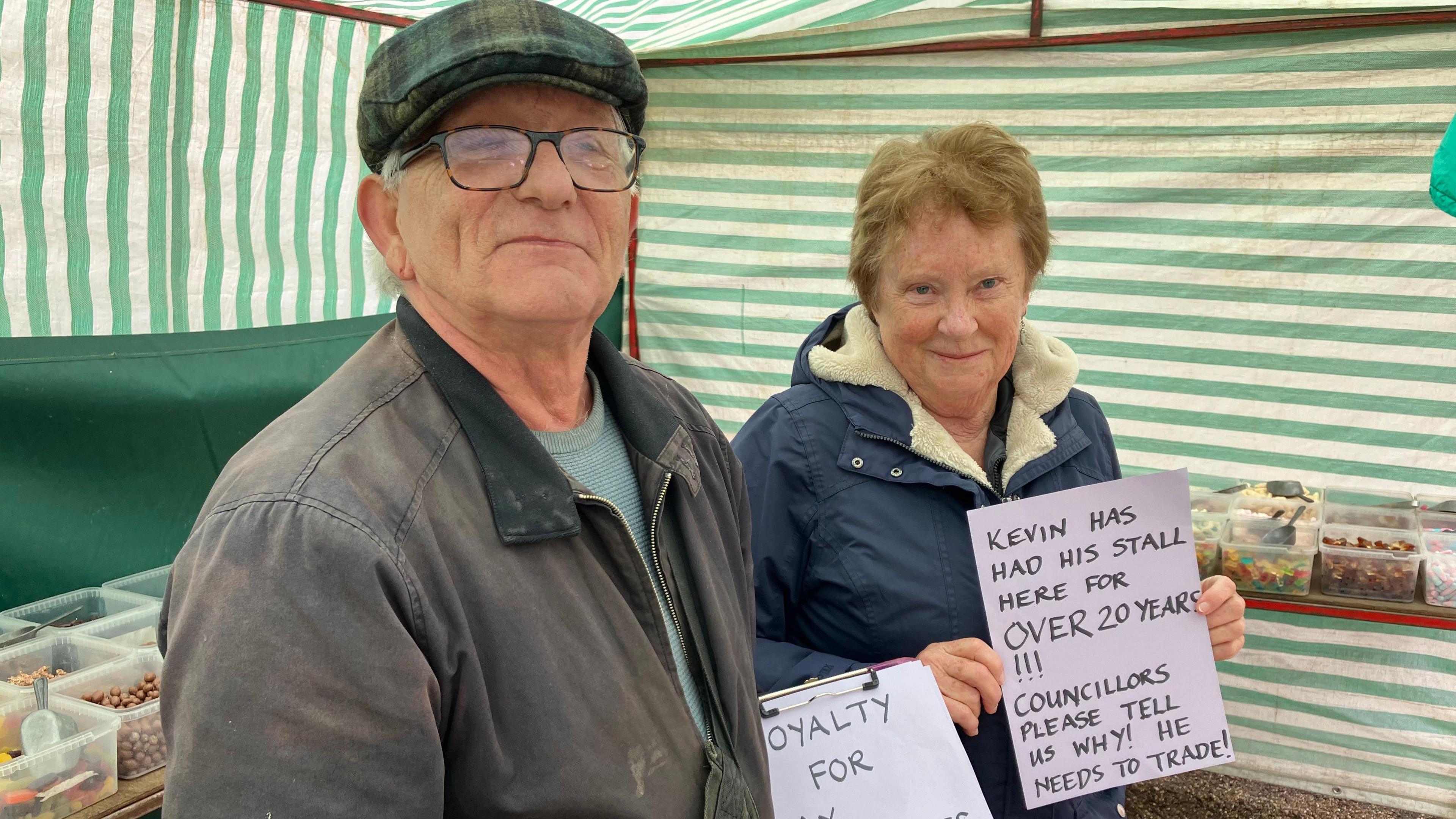 Stall holder wearing cap standing under market awning with a female supporter 