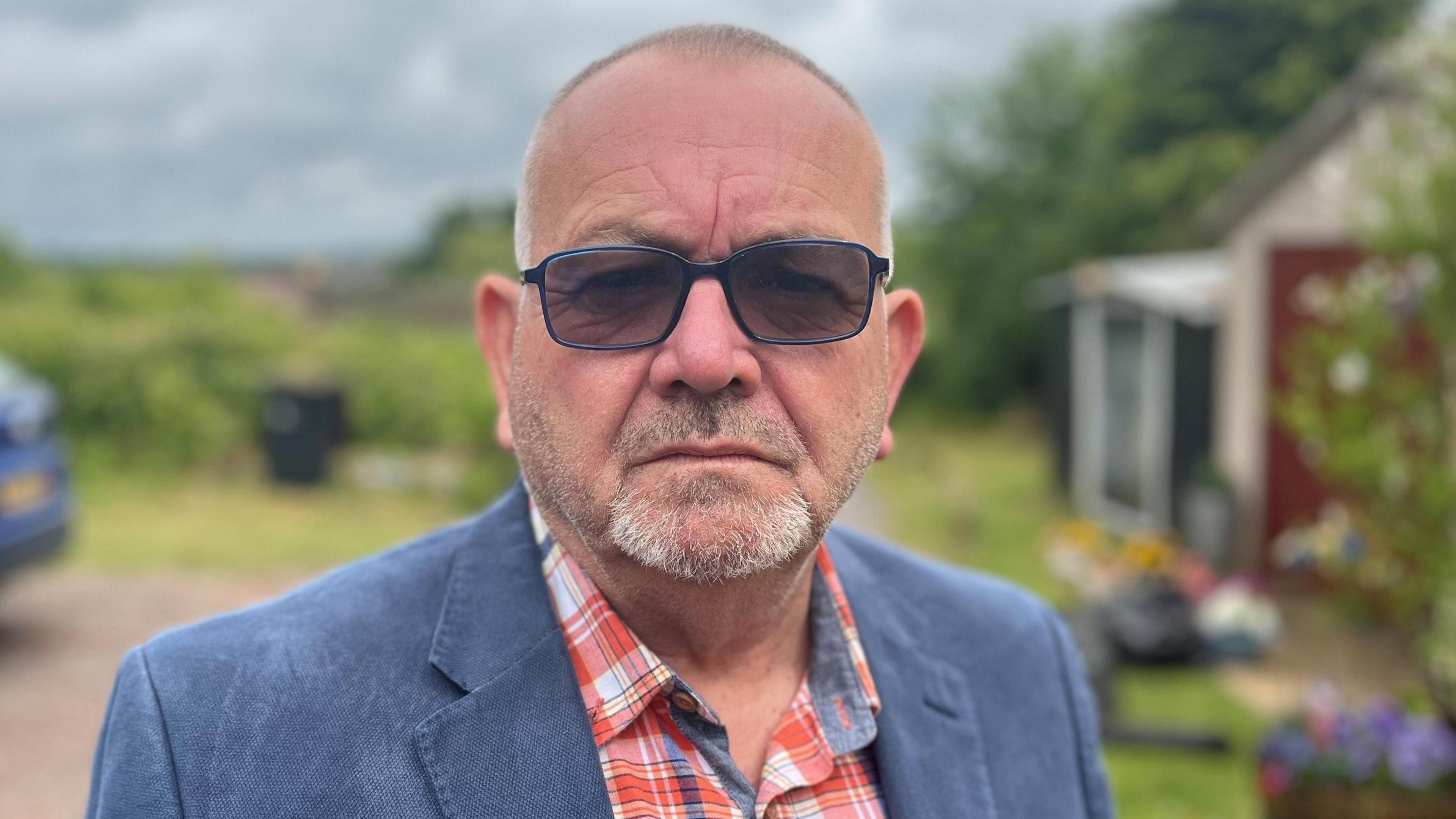 Martyn Underhill has short, grey hair and a short beard. He wears sunglasses, a blue jacket and a checked, red shirt.