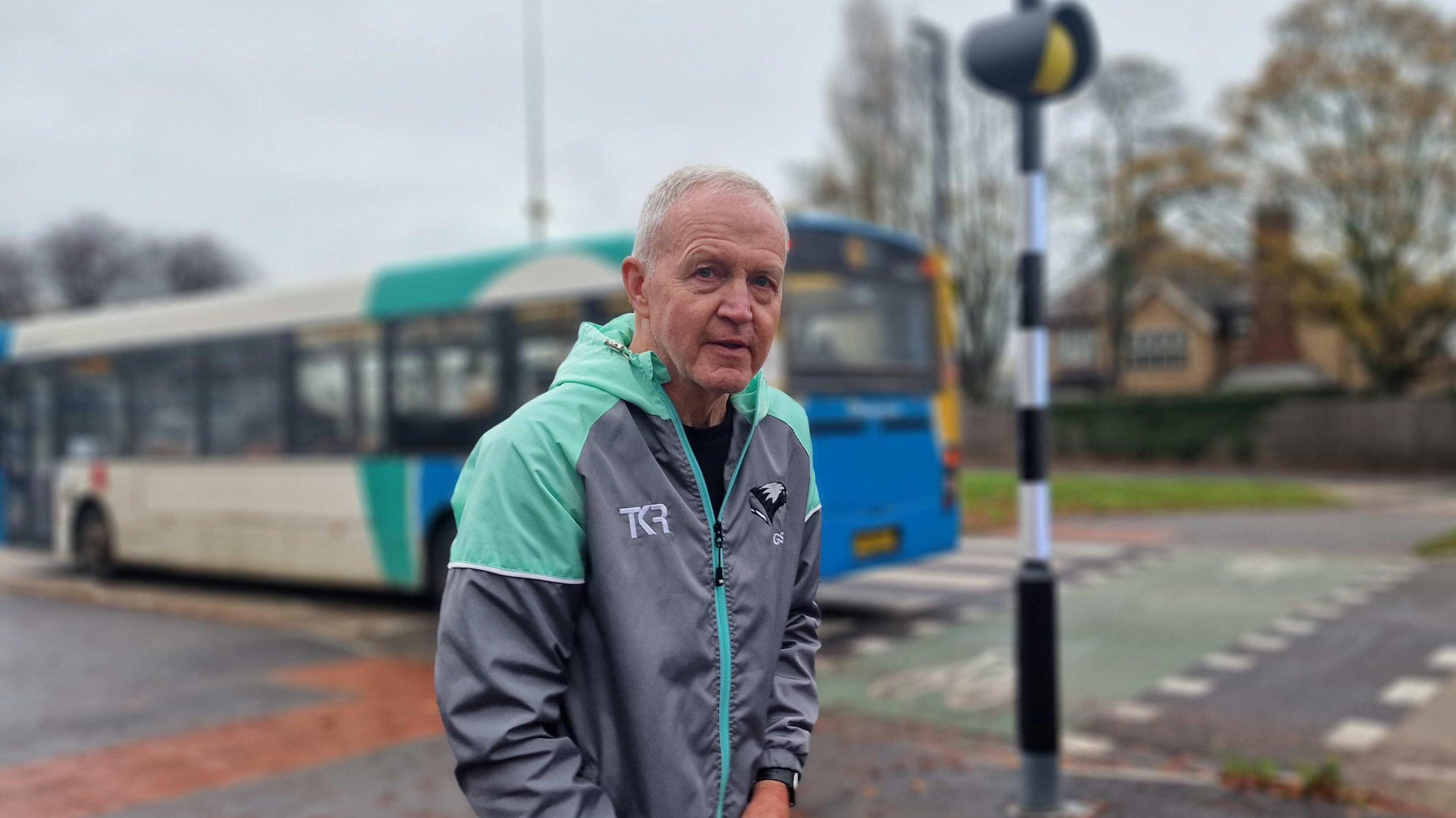 Man with short hair and blue and grey jacket. There is a bus behind him, a zebra crossing, a residential building and trees.