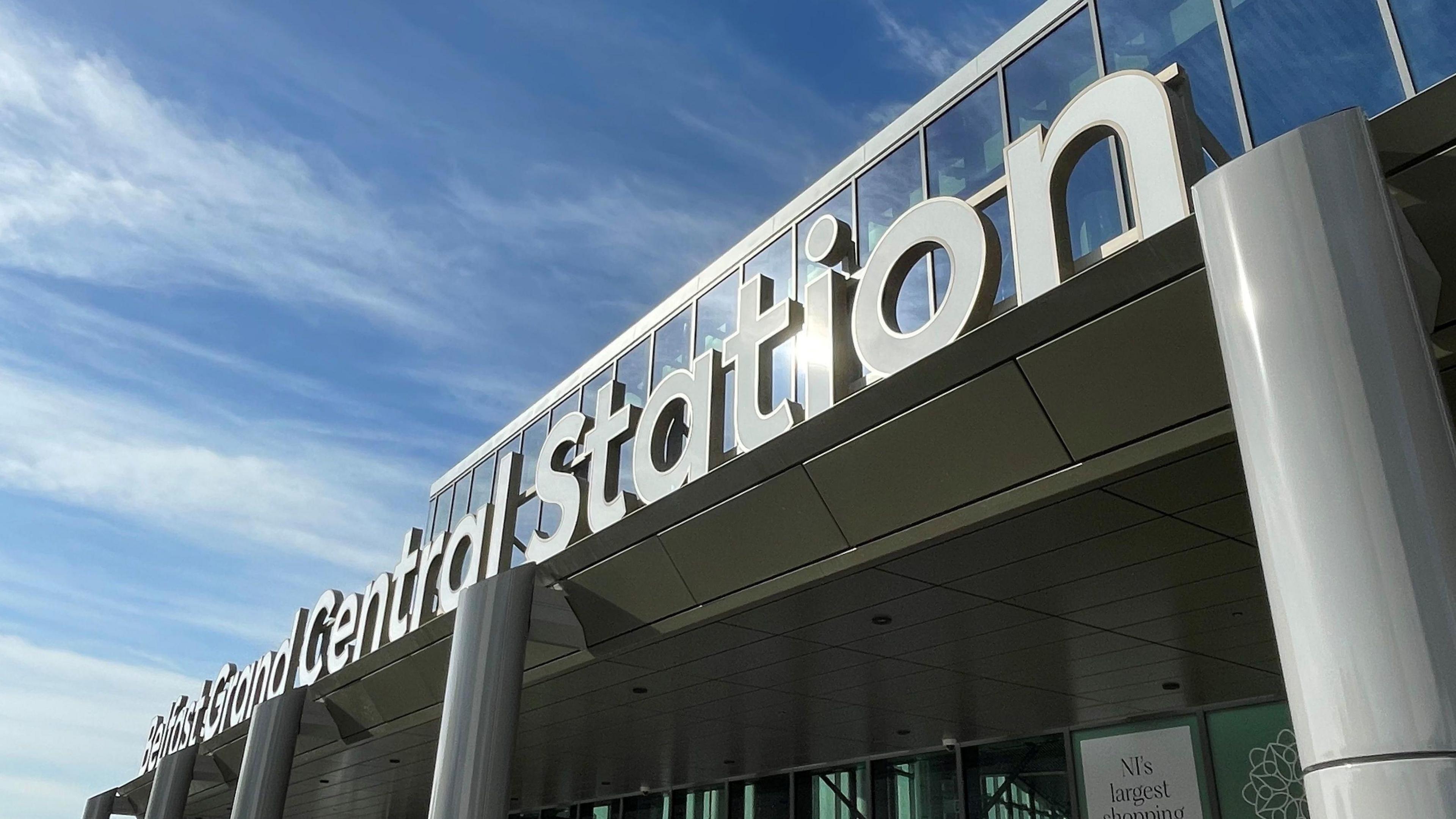 The front entrance to Belfast Grand Central Station. The building is silver metal and there is a sign in silver metal that reads 'Belfast Grand Central Station', the sun is shining onto the building causing a shimmer on the metal.