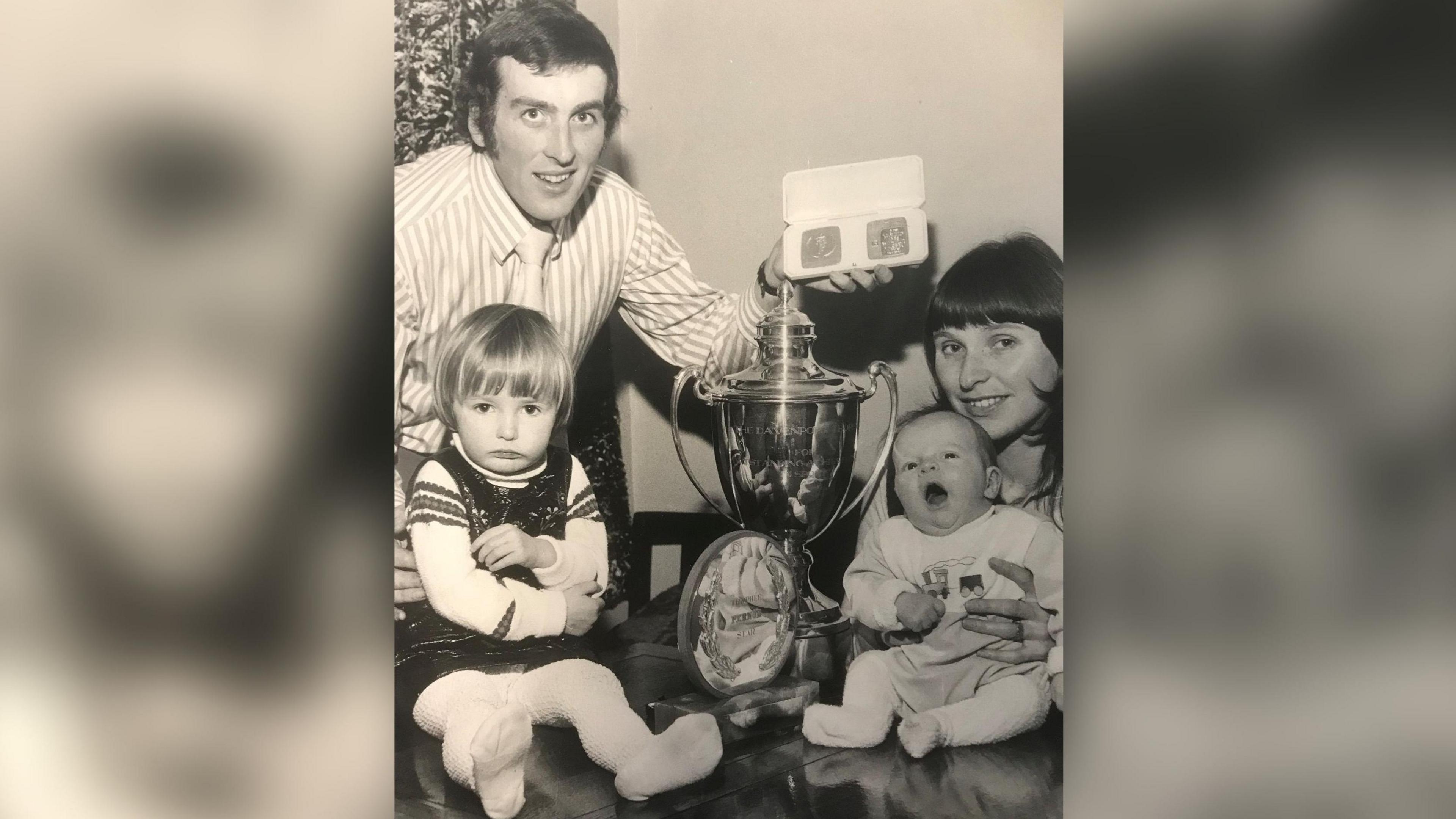 A man and woman and a little girl and a baby are pictured in a black and white photo. The man is holding medals and there are two trophies in the middle of them all.