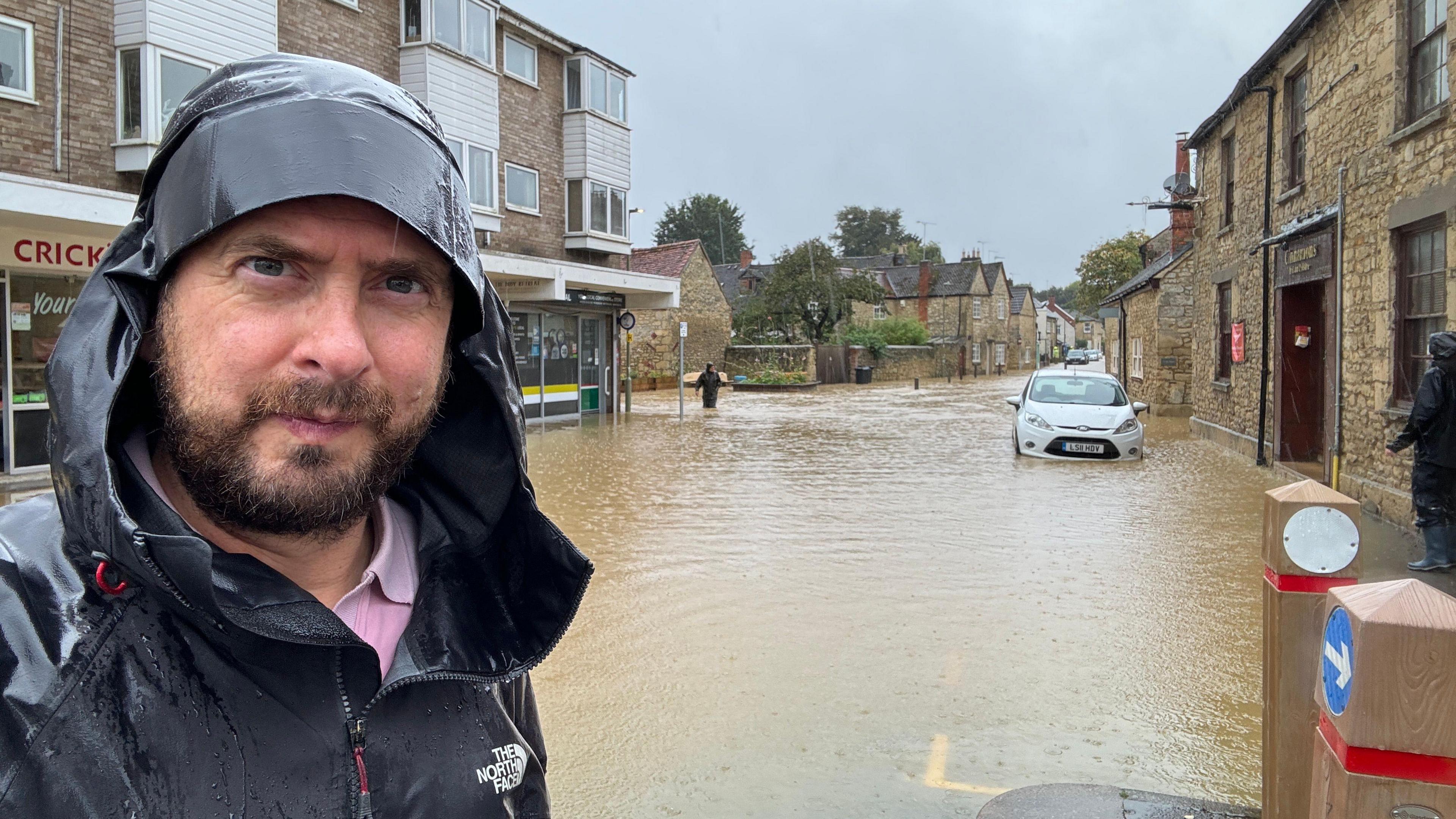 Tim Bearder is wearing black coat with his hood up. Behind him is a street submerged in brown murky water.