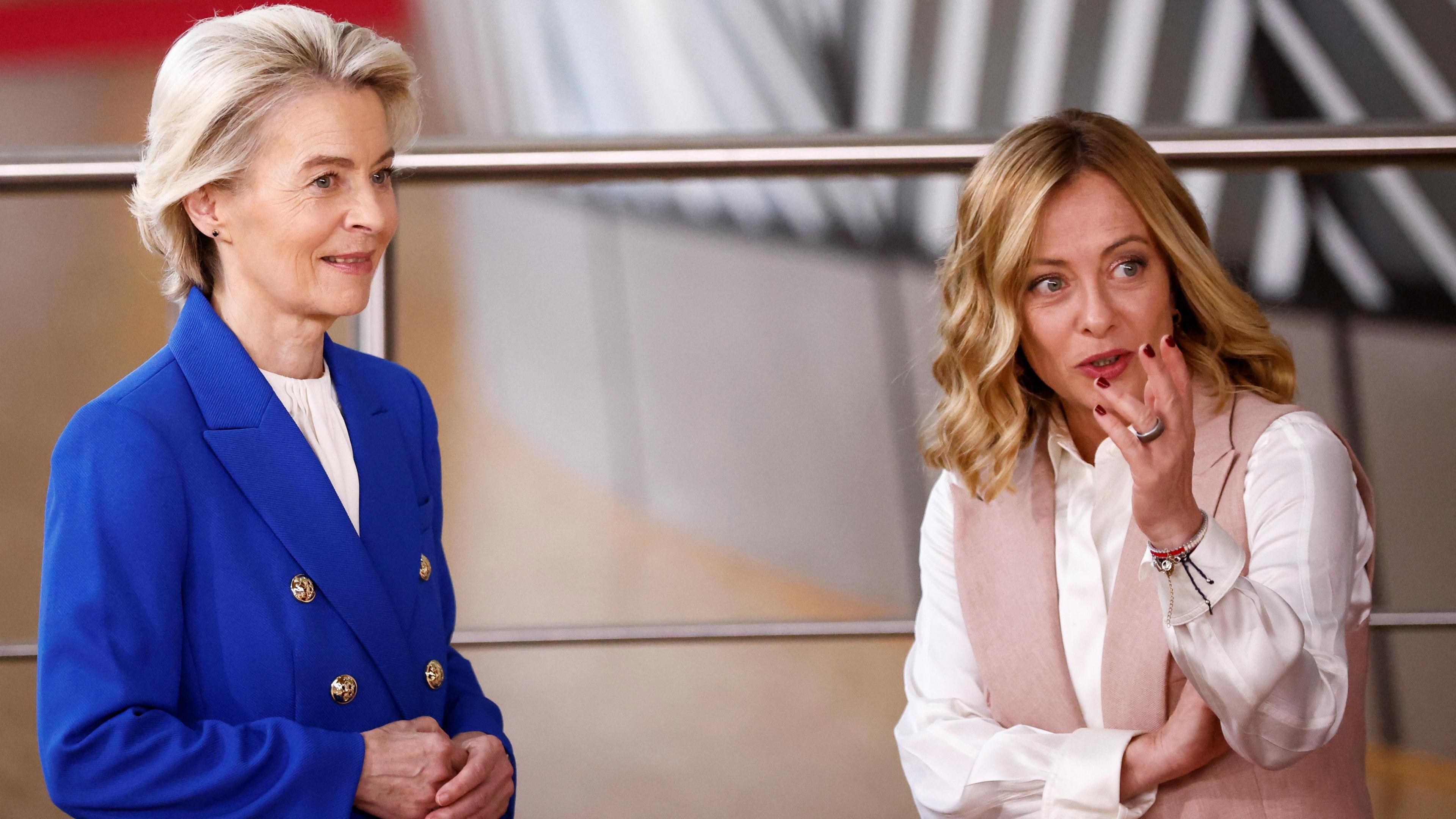 Ursula Von der Leyen, with a distinctive short blonde haircut, wearing a royal blue jacket with gold buttons and a white blouse, reminiscent of the EU flag, and Giorgia Meloni, who has blonde hair which has been curled, wearing a pale pink waistcoat and gesturing with her left hand. They are both looking off camera.  