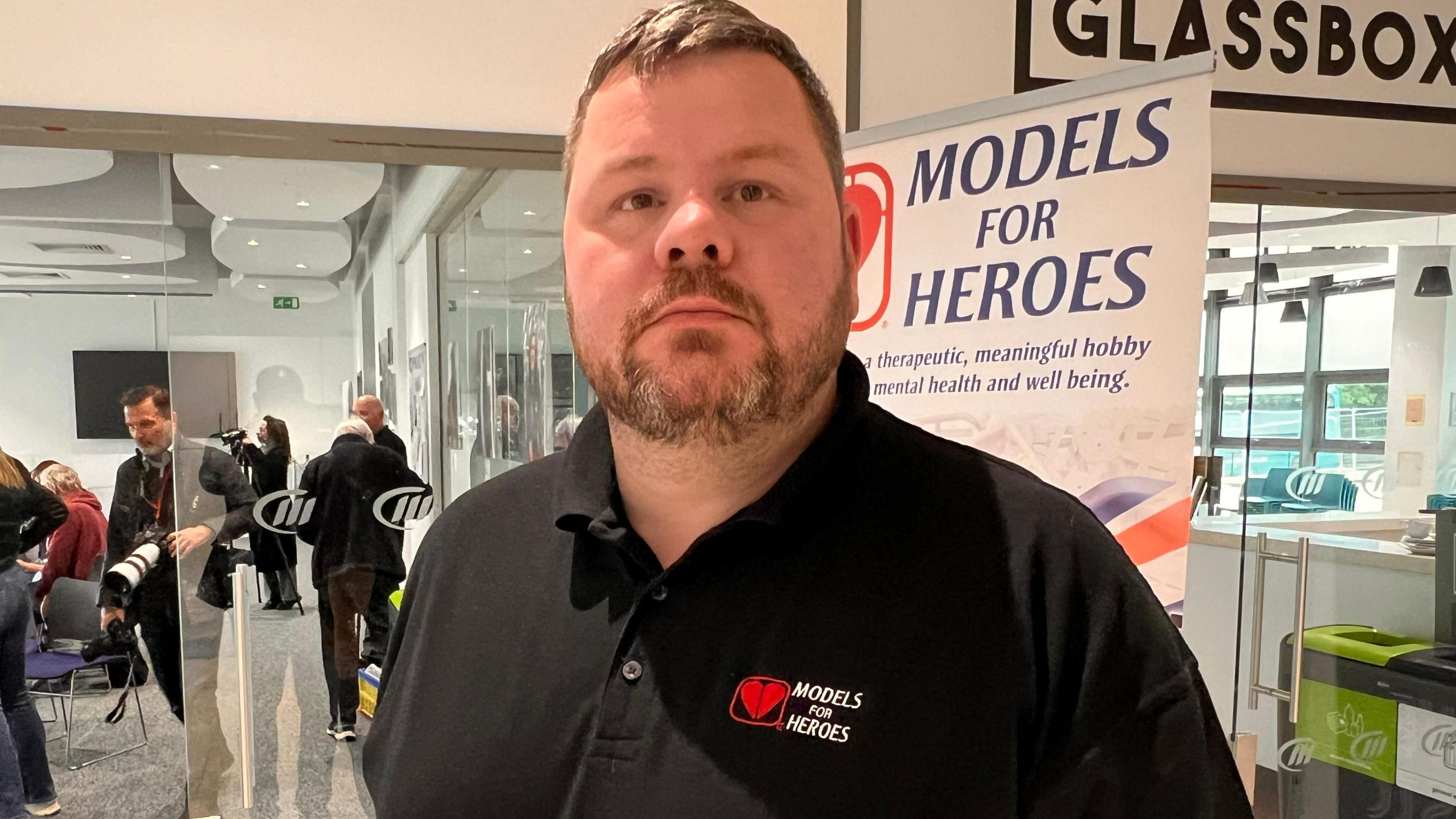 A man wearing a black t-shirt bearing the charity name Models for Heroes is looking directly at the camera