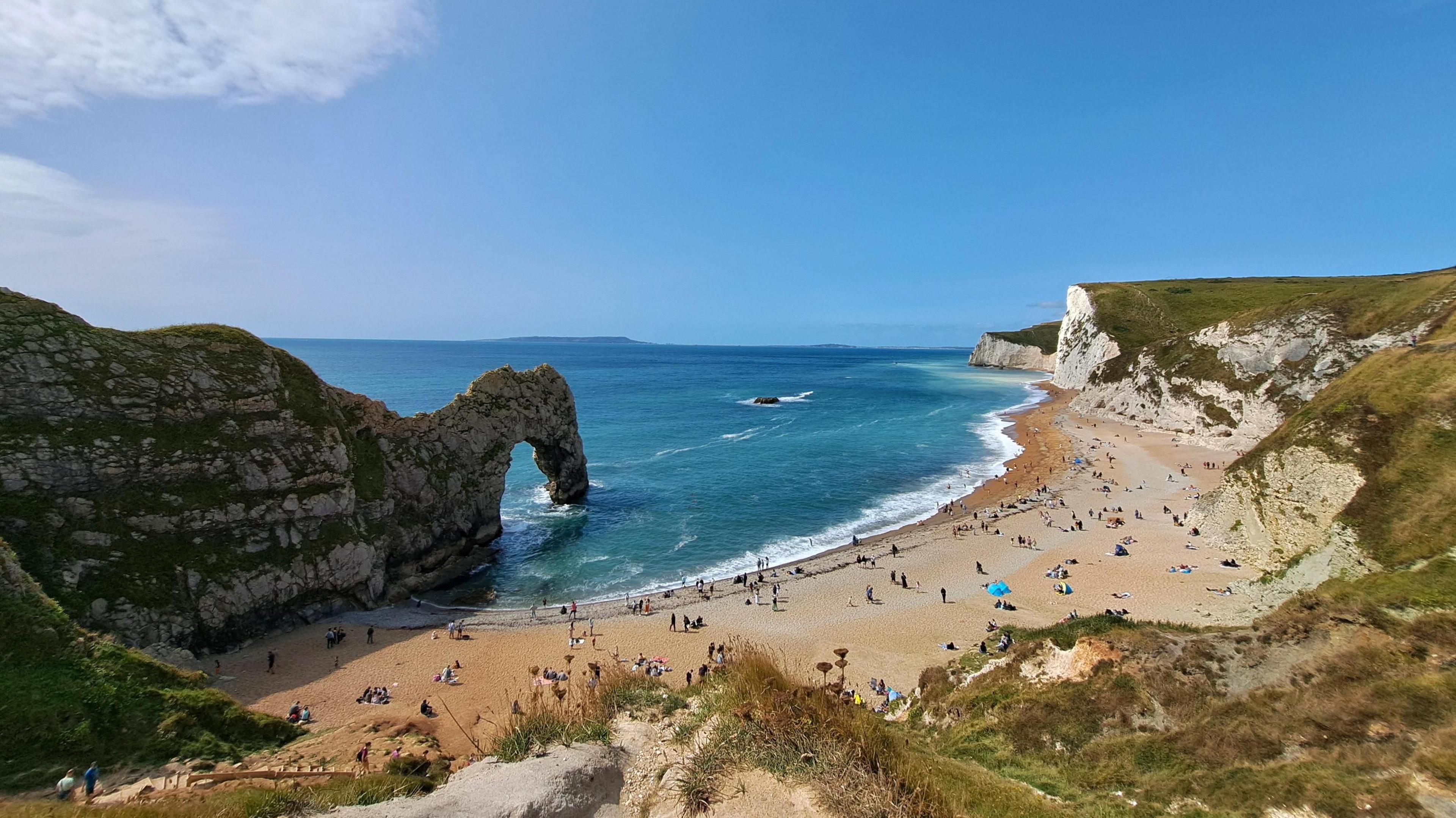 The rock arch at Durdle Door with the cliff behind on a sunny day. There is a sandy cove with hundreds of people sitting on the beach in front of the cliffs. The cliffs are covered in green grass and the sea is clear blue. Overhead the sunny sky is blue with one white cloud.