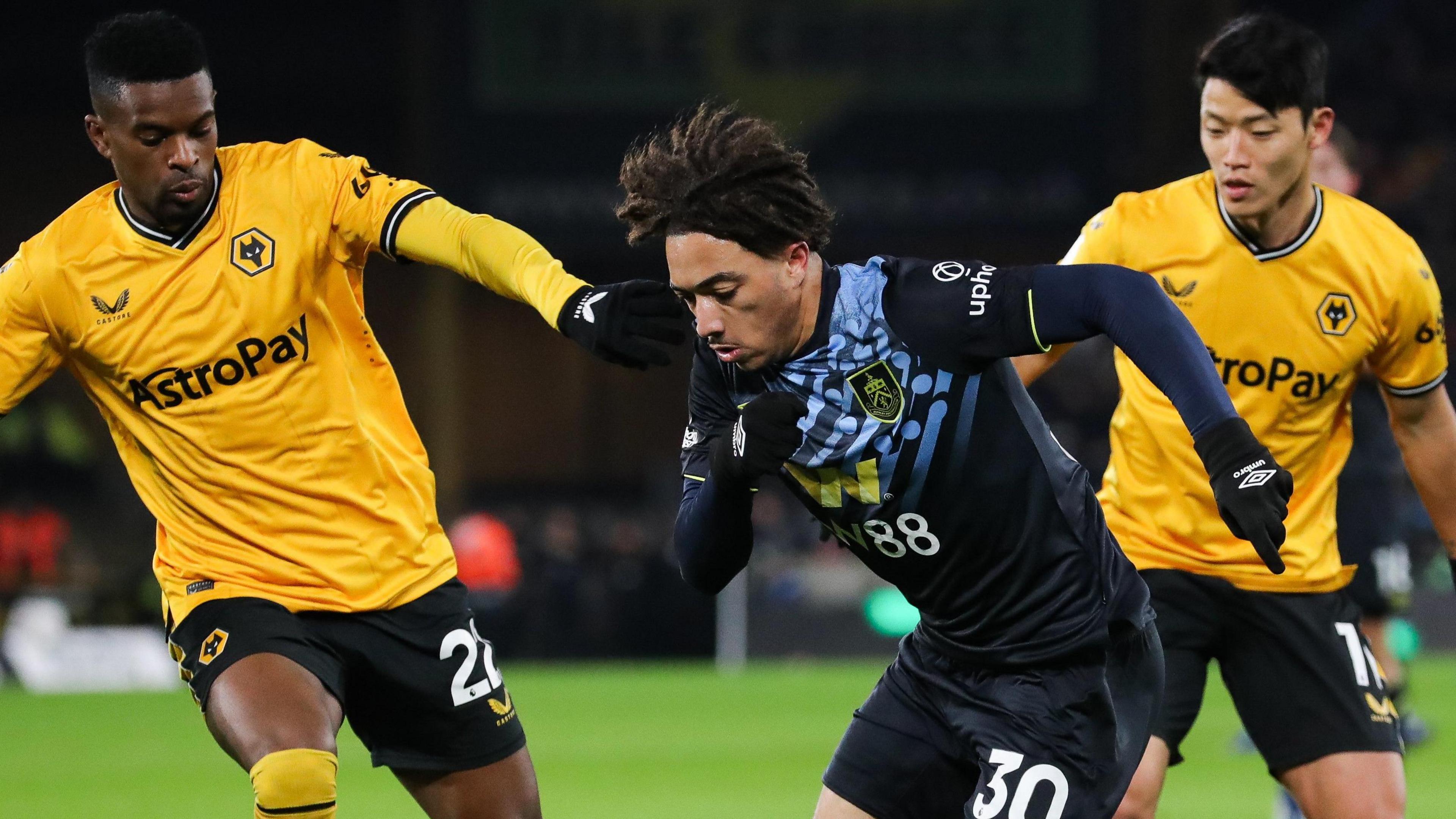 Luca Koleosho, wearing Burnley's black away kit, runs past Wolves' Nelson Semedo on his right. Semedo's right arm is stretching out to Koleosho's shoulder as he looks down at the ball on the ground (not in the picture) 