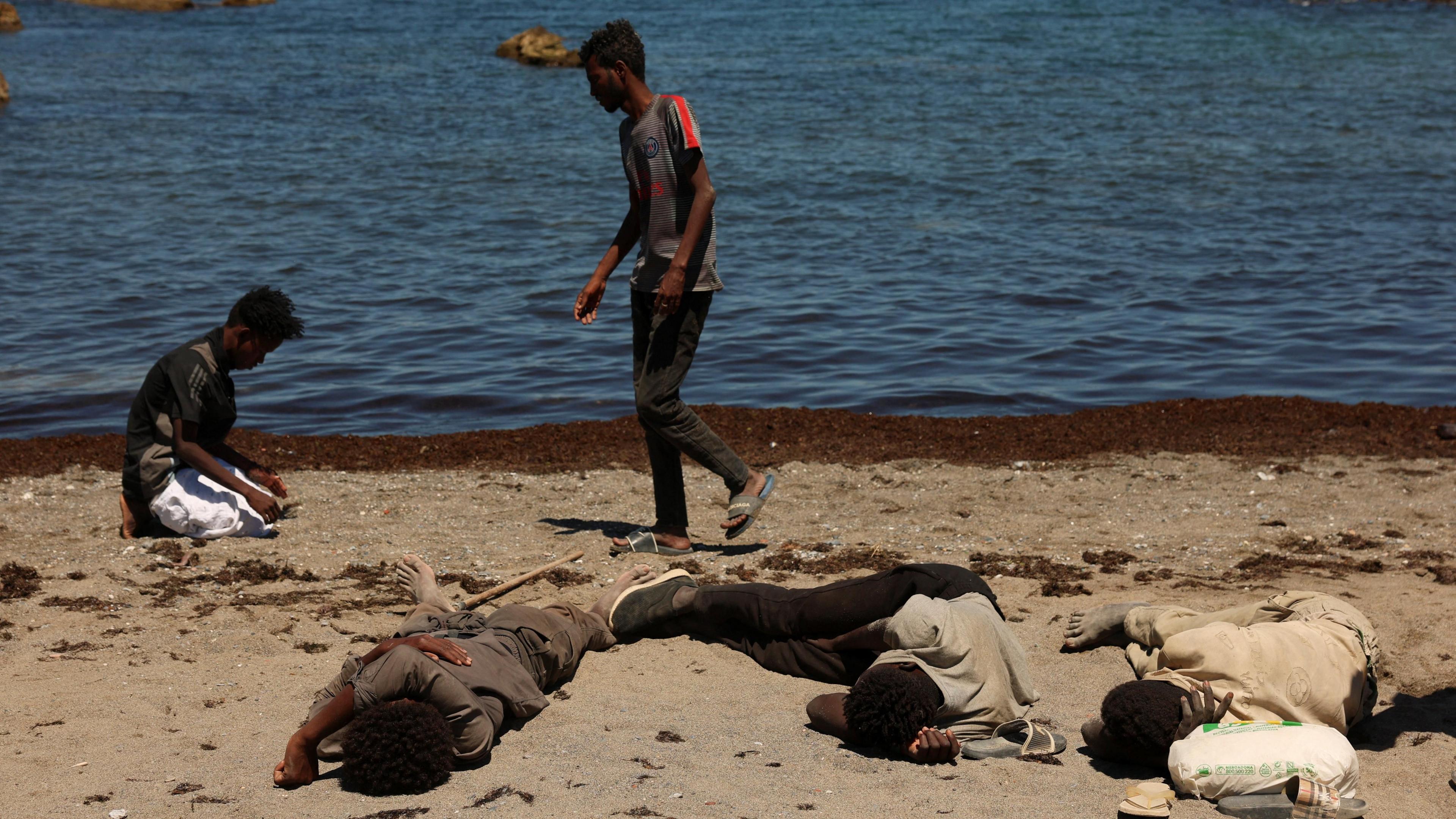 Three men sleep on a beach while another prays in front of the sea in Ceuta