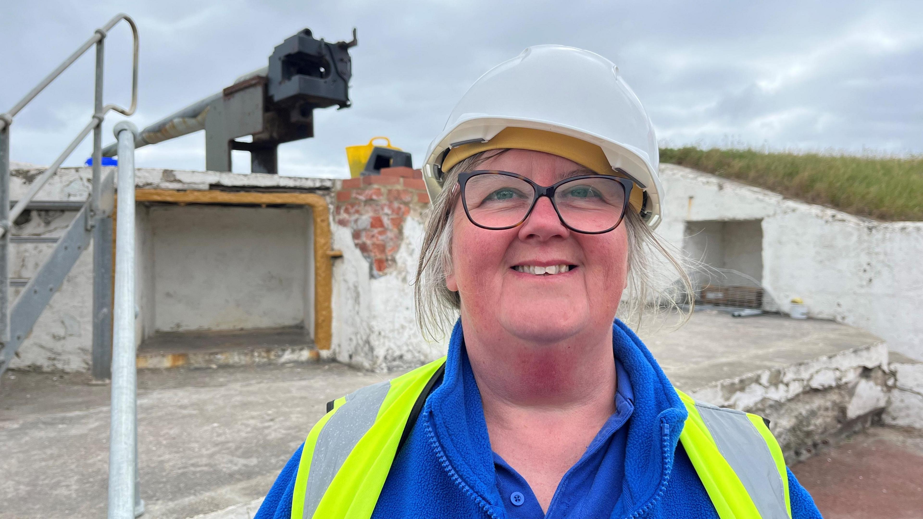 Diane Stephens, a blonde-haired woman stands in front of the camera smiling, wearing a white hard hat, blue fleece and fluorescent tabard.
Behind her is a World War One gun battery.