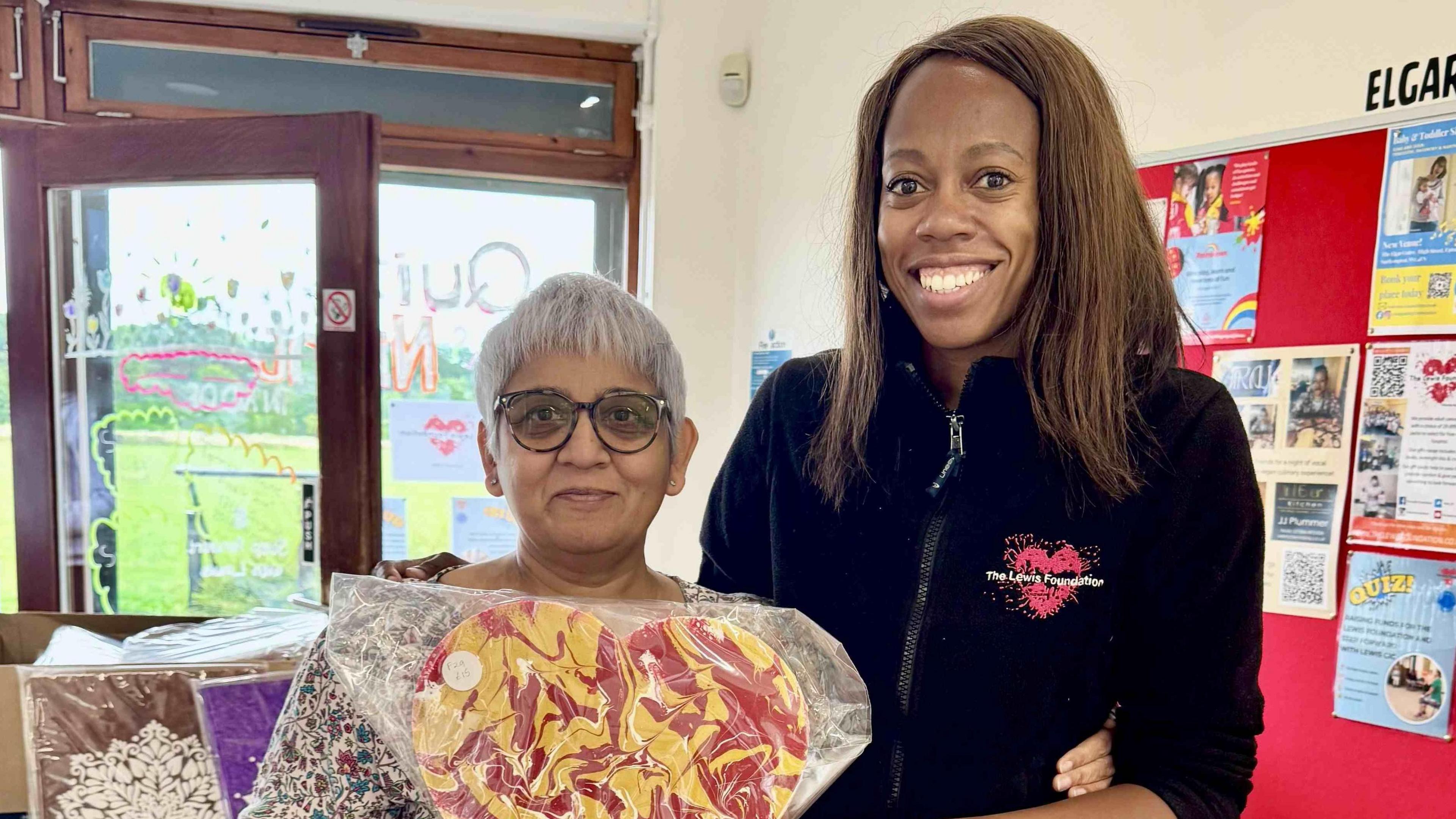 Jagruti Patel and Lorraine Lewis pose for a picture inside a community centre. An open playing field is visible out of double doors behind them.