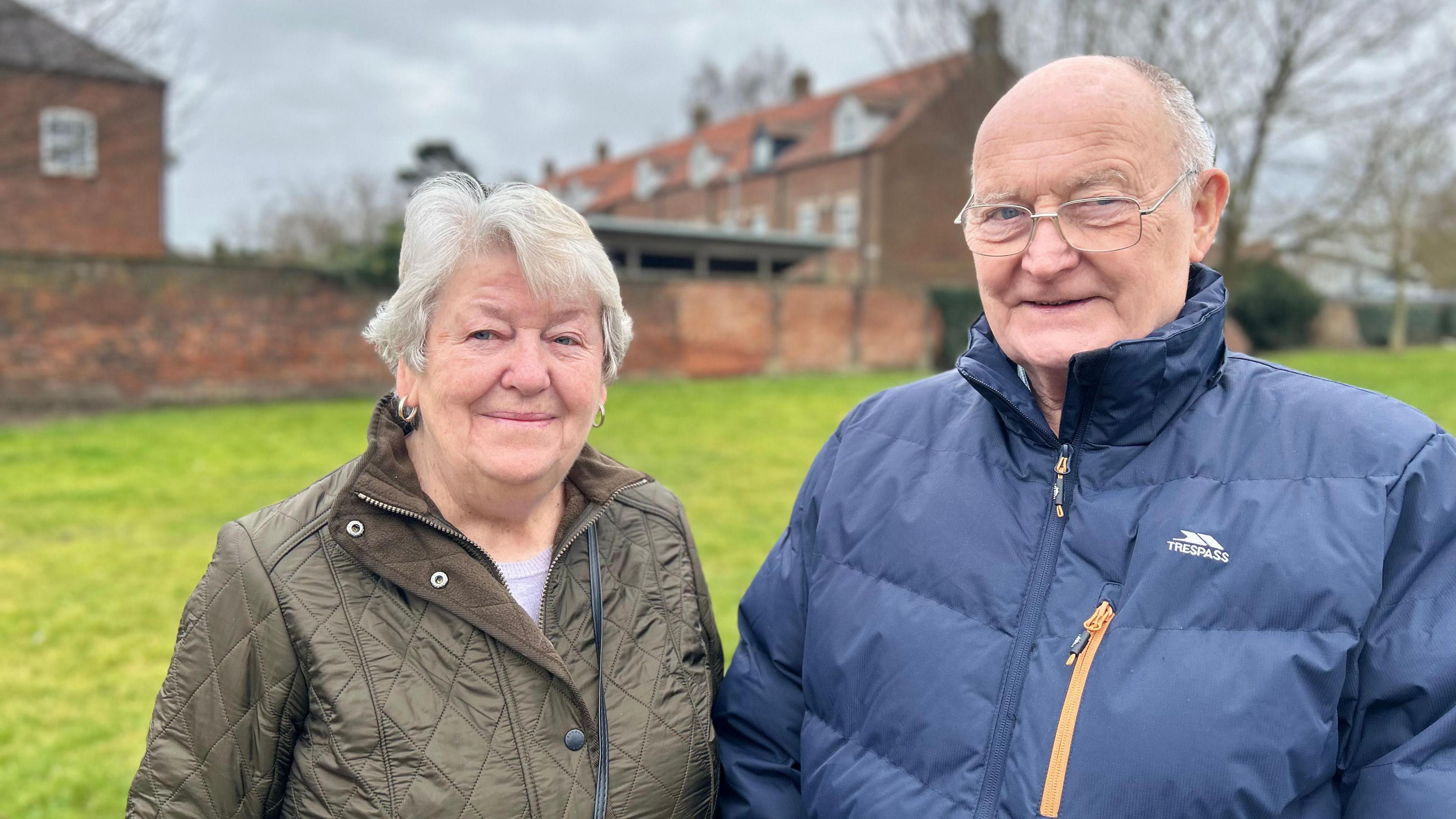 A couple, both dressed in winter coats, stand on a roadside in front of a green open space, with red-brick terraces in the background. 