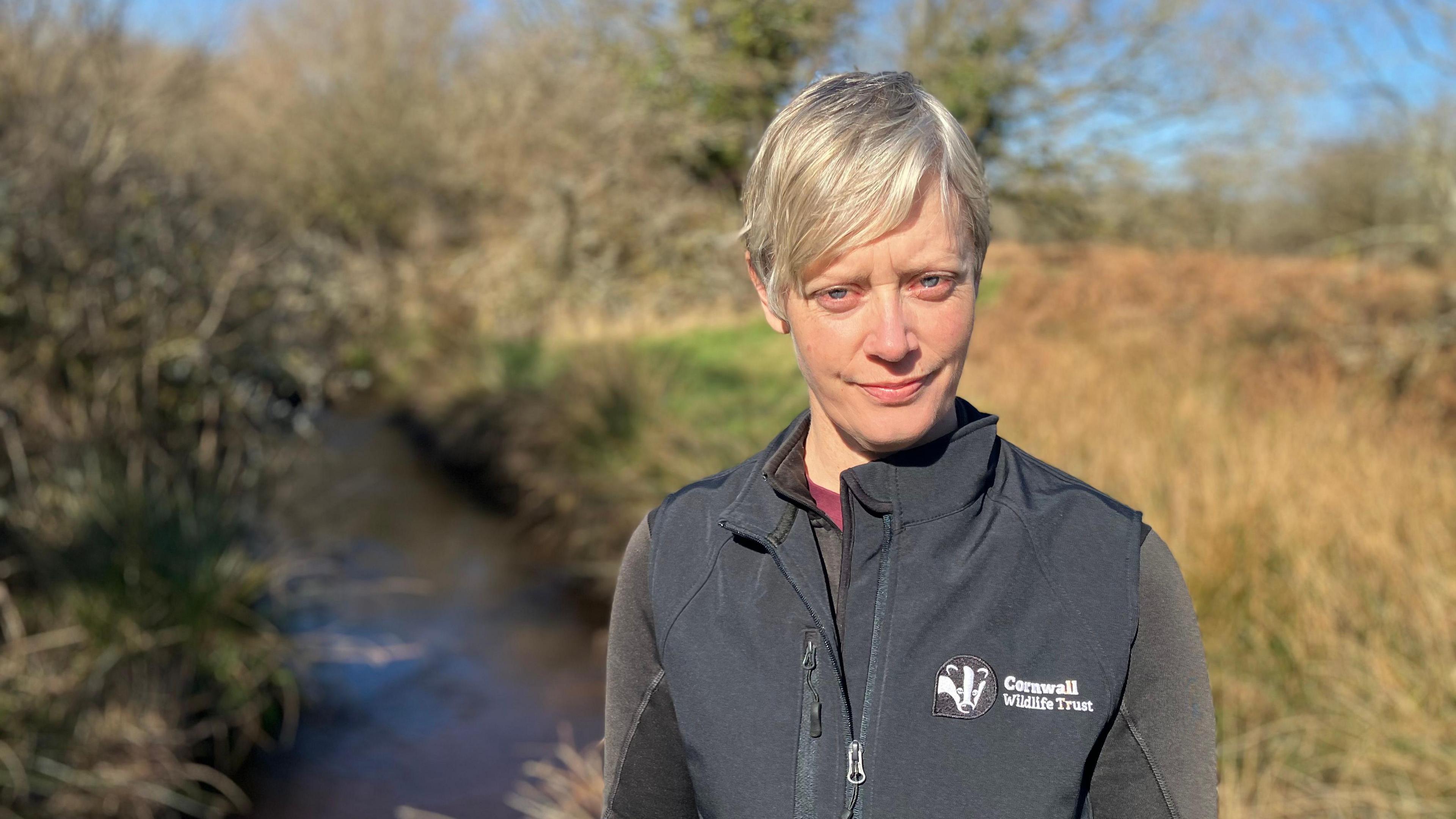 A woman stands in front of a small stream, there is grass and shrubs in the background. She is wearing a black gilet with the Cornwall Wildlife Trust logo on it. 