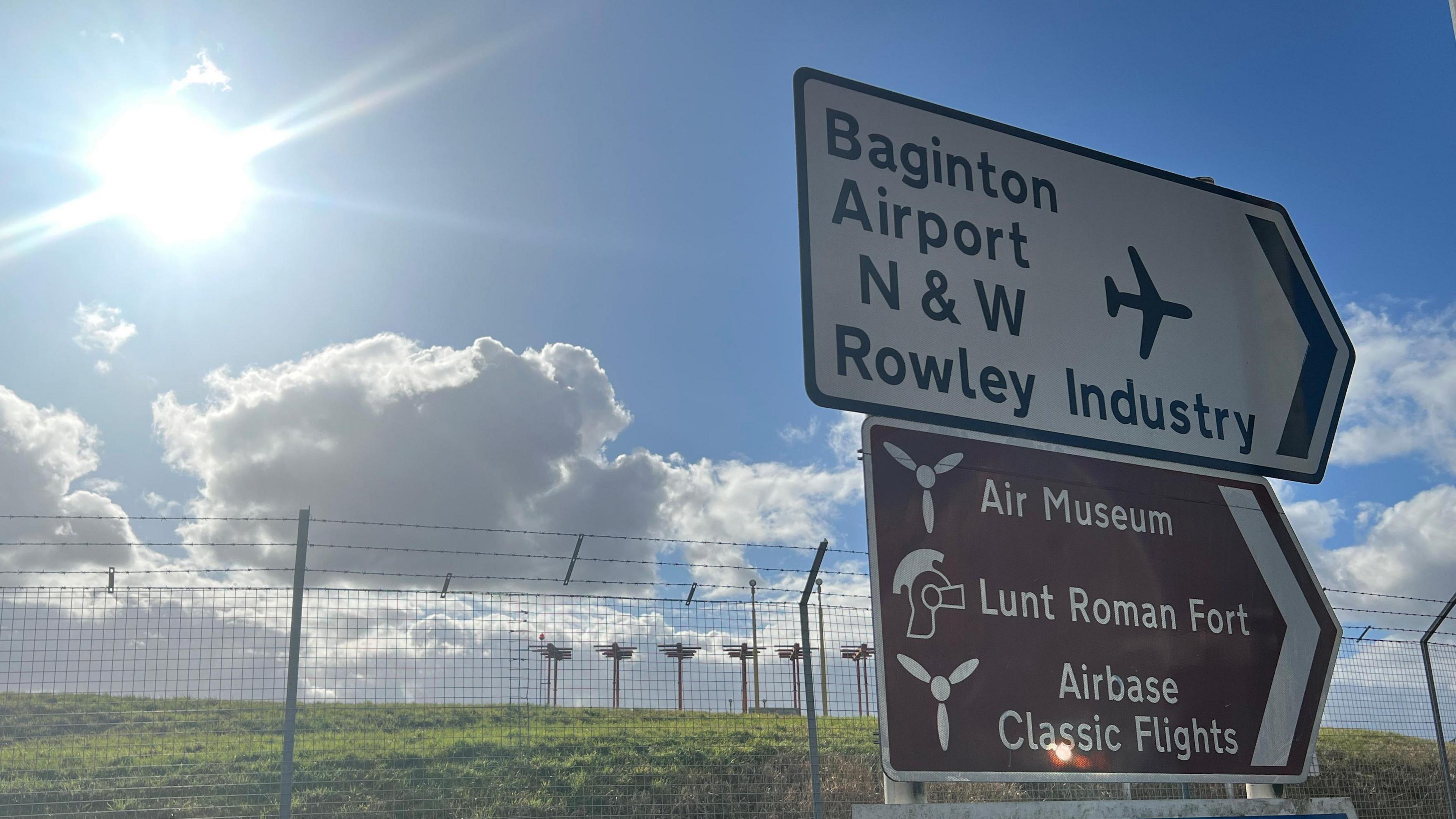 The sun shines above the airport runway at Coventry Airport. In the foreground we see a road sign which reads "Baginton Airport" with the airport barbed wire fence behind.