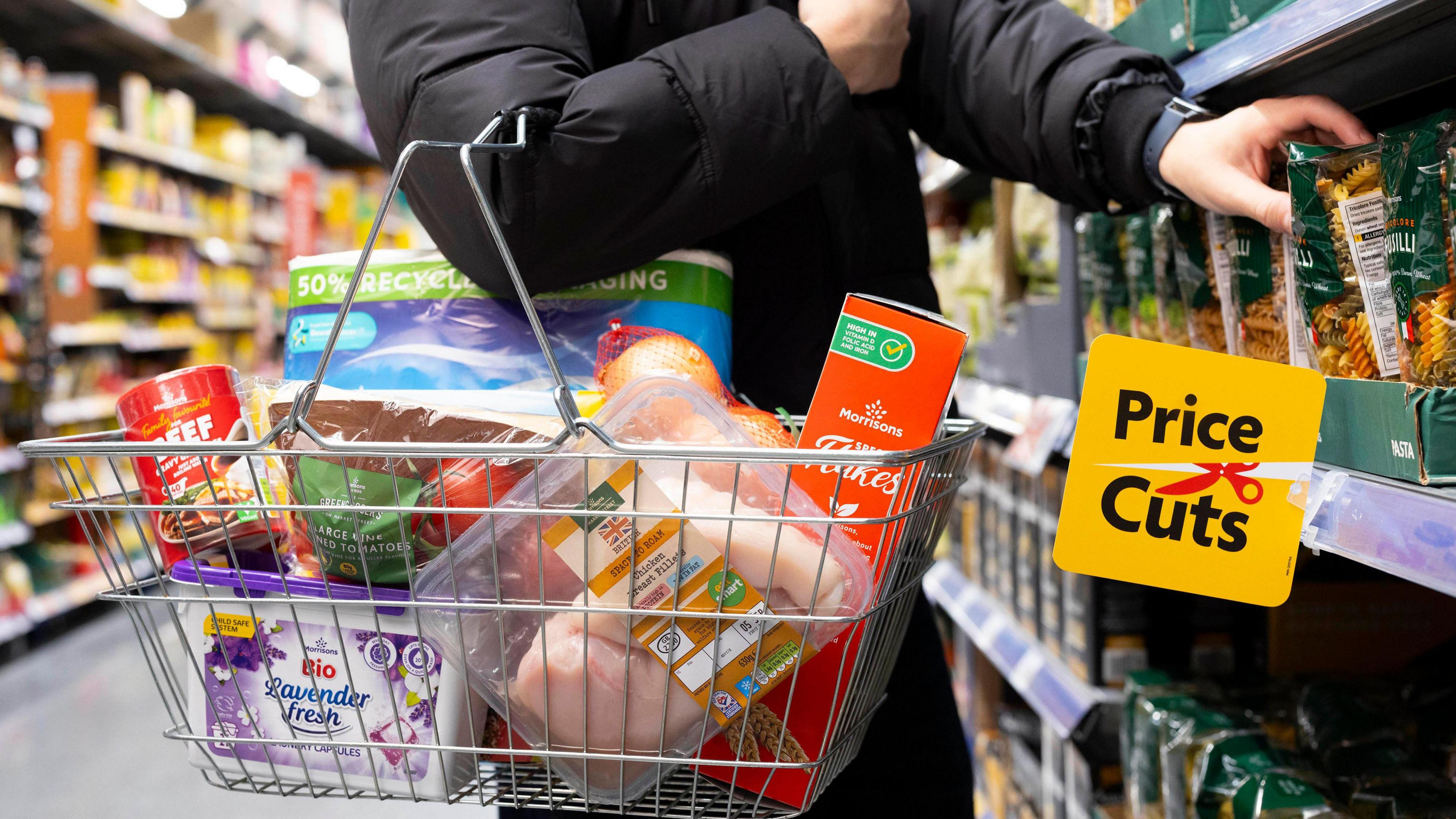 A person wearing a black coat in a supermarket aisle taking pasta off the shelf to put it in their very full basket. A yellow sign which reads reads Price Cuts next to a red pair of scissors hangs fro the shelf.