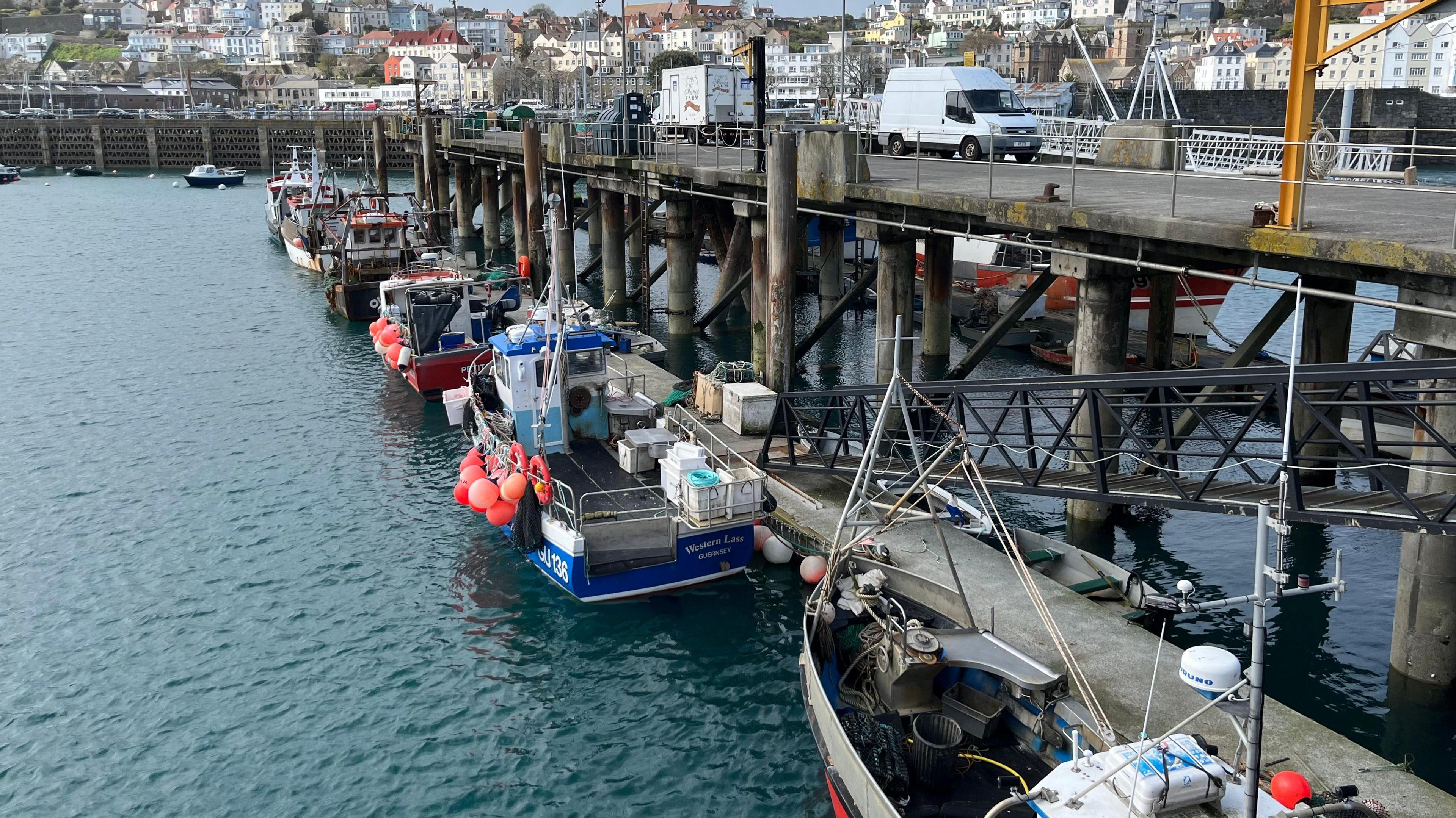 Guernsey's Fish Quay. Five fishing boats are docked next to a grey pier with commercial fishing equipment visible on board.