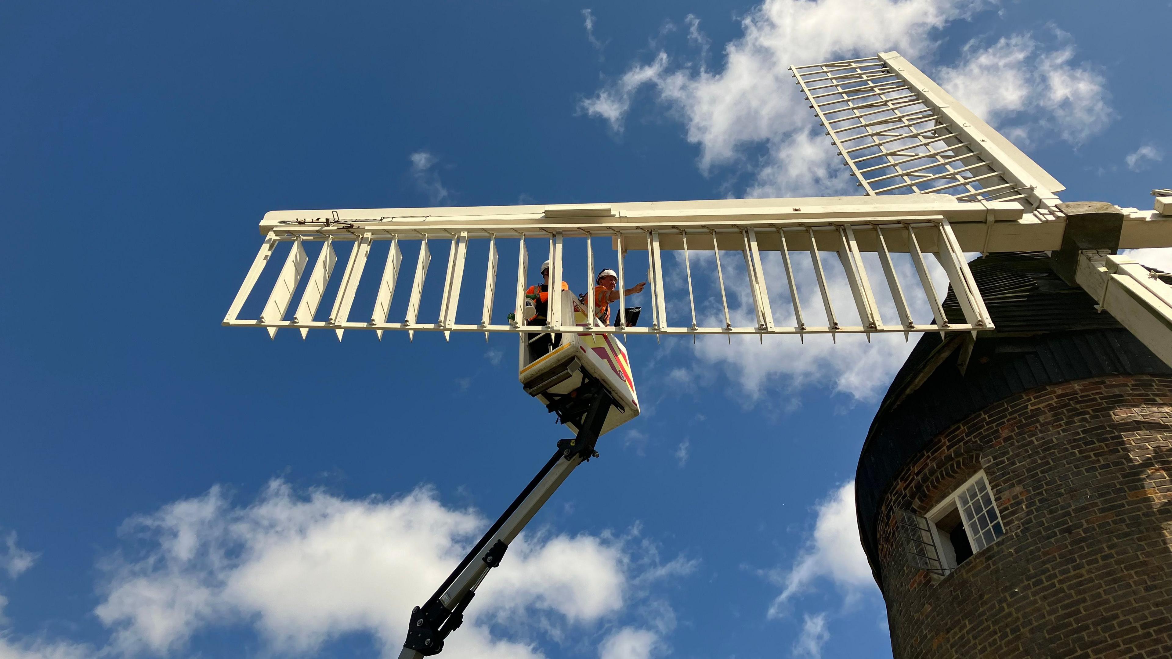 Two volunteers stand on a platform to repaint windmill sails