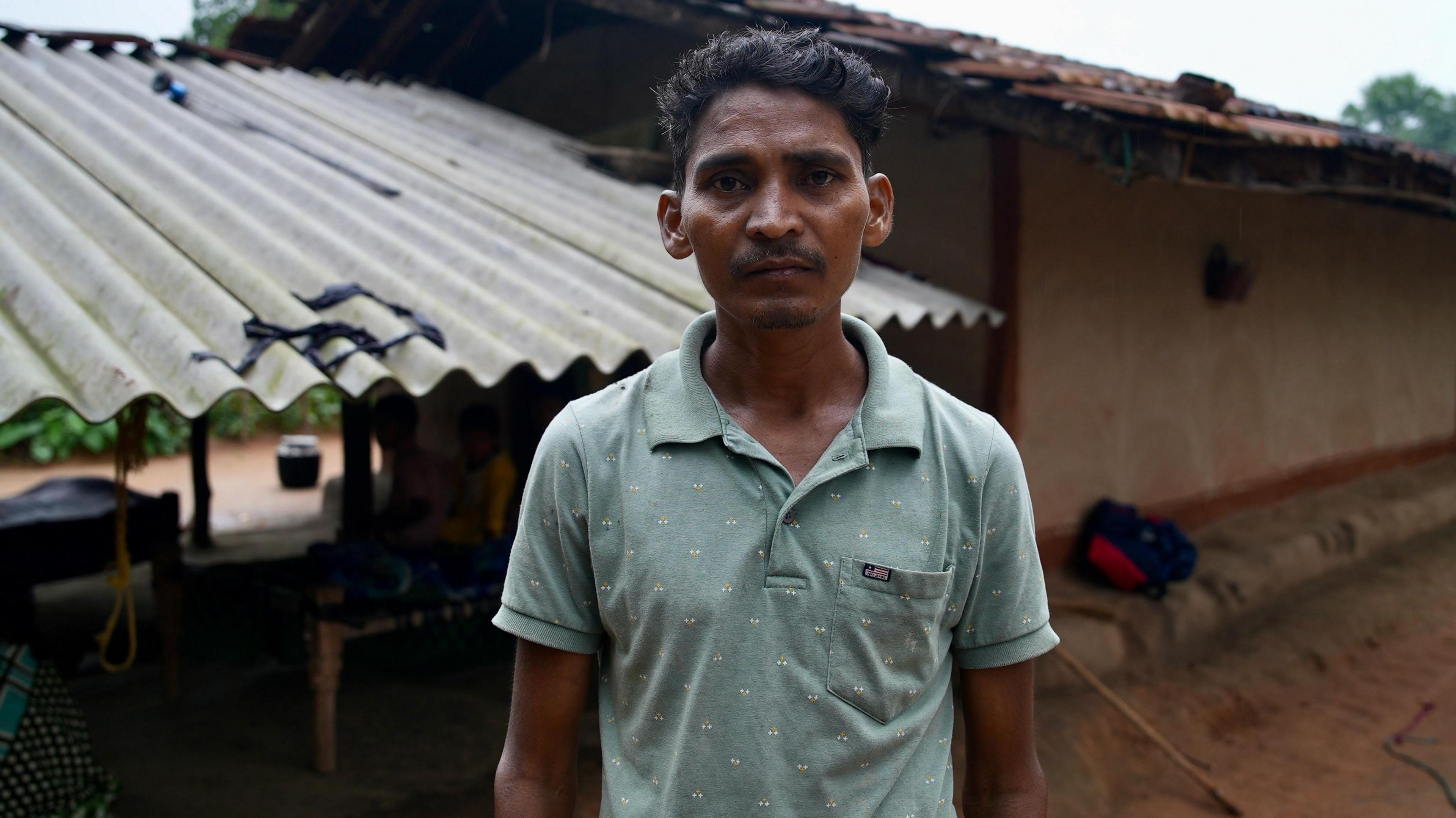 A man wearing a blue tshirt looks into the camera. He is standing in front of a shanty house. 