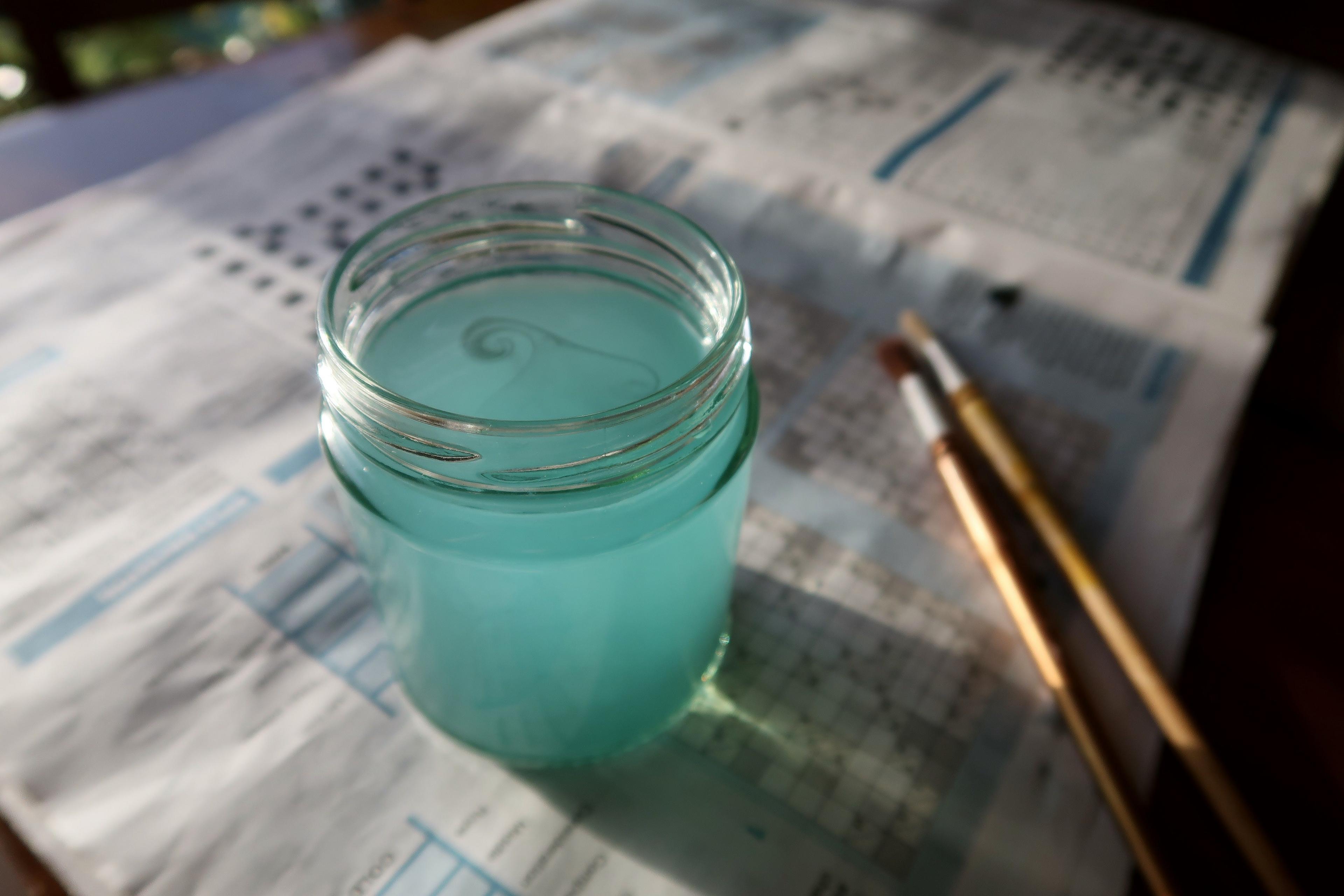 A jar with green water rests on newspaper with paint brushes nearby