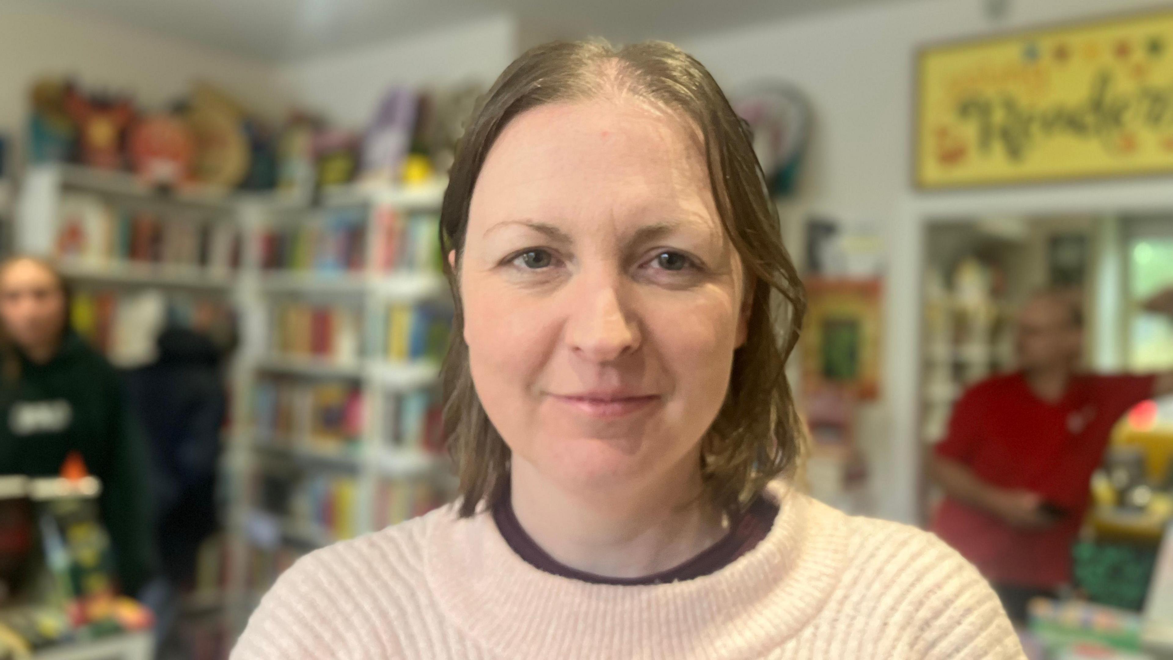 A woman in a pale pink jumper stands in a book shop. She has brunette hair. Behind her book shelves are blurred.