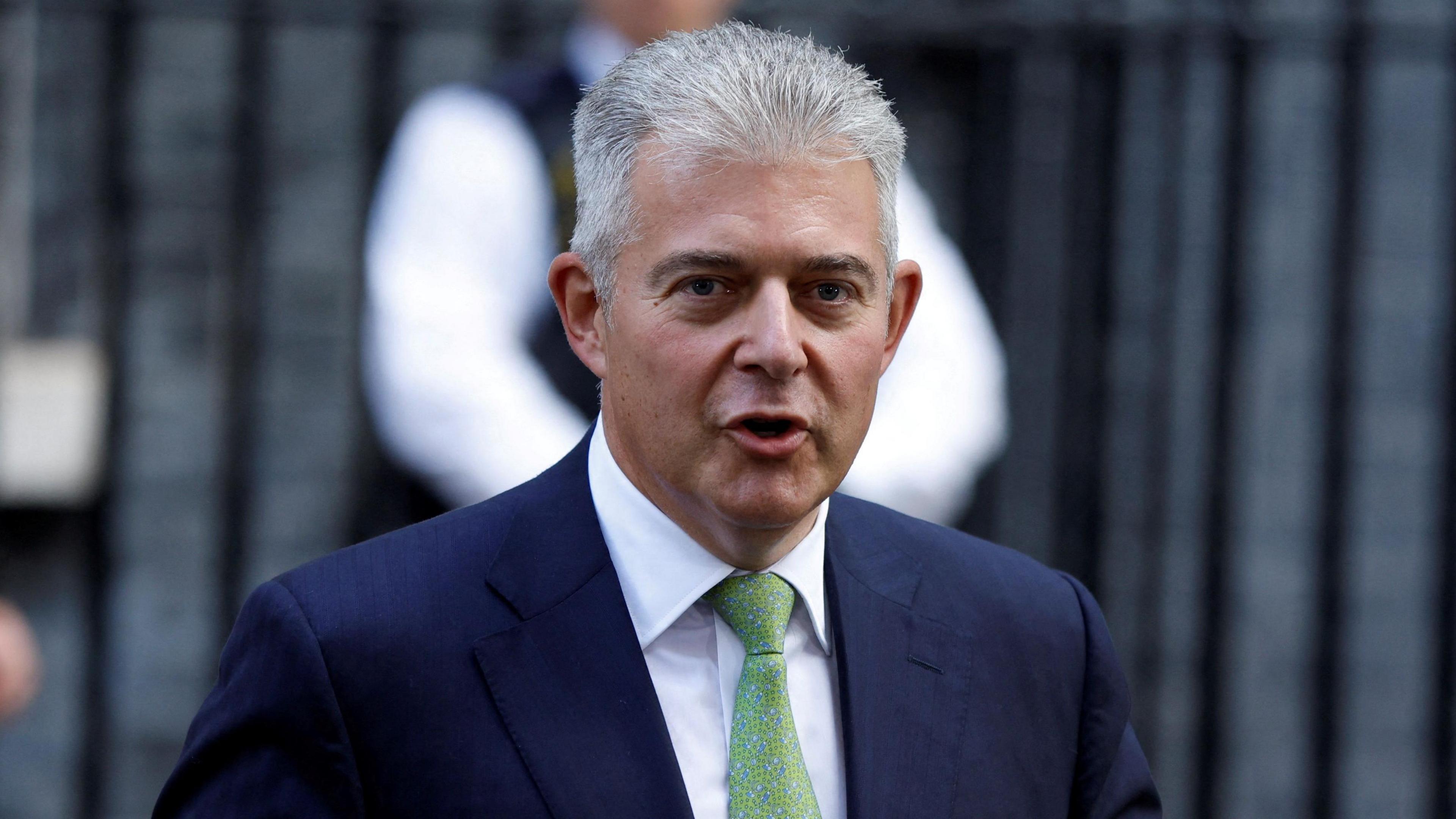 Brandon Lewis walking down Dowing Street carrying a folder under his arm. He is looking at the camera and saying something. He is wearing a suit and tie. Behind him out of focus is a man in police uniform and and another man wearing a suit and carrying a folder. Behind them the black railings and bricks of Downing Street buildings.