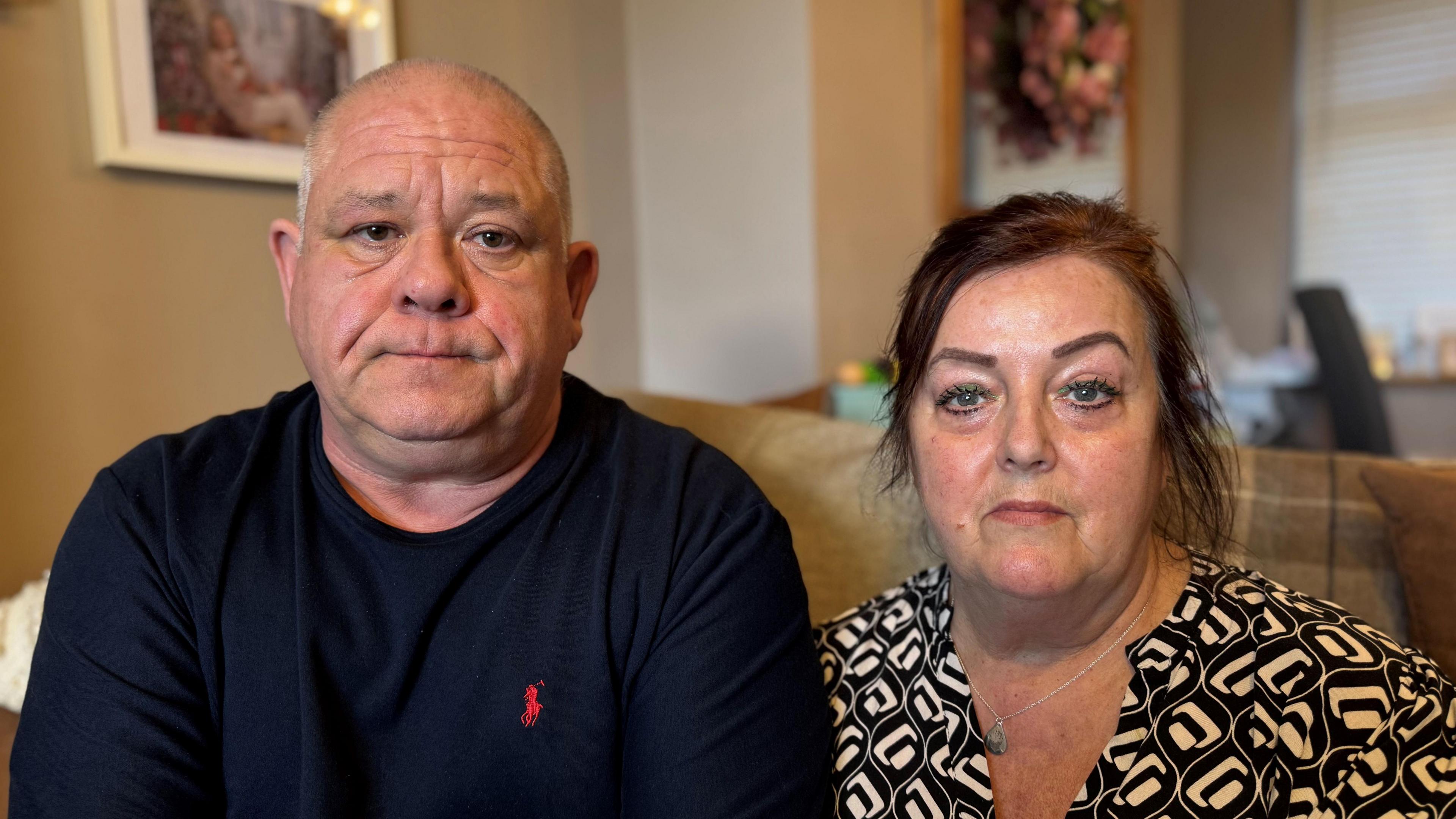 Elizabeth and Jonathan look at the camera. They are sat on a sofa, Jonathan has a shaved head is wearing a dark top with a red Ralph Lauren polo logo, and Elizabeth has dark hair which is tied up and is wearing a black and white patterned top. 