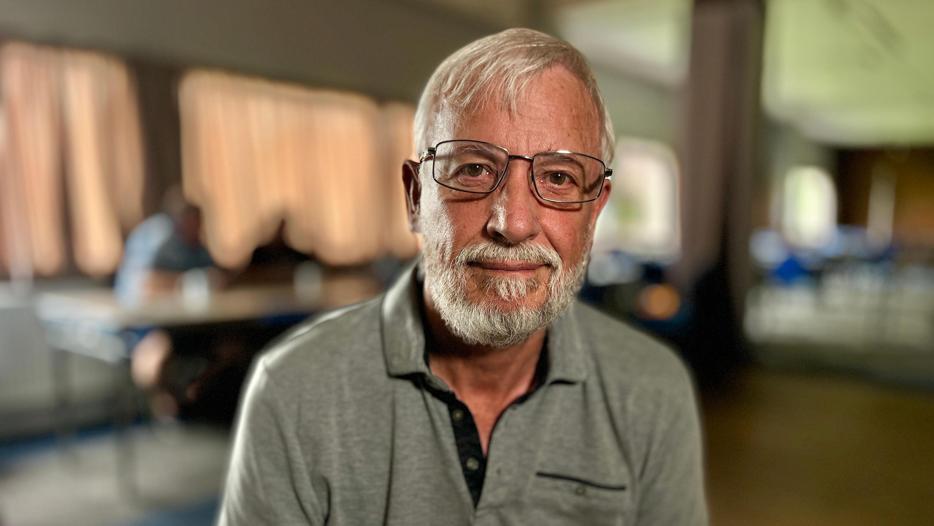 Jim Smith looks at the camera. He is wearing glasses and has a grey beard. He is wearing a grey polo shirt. The light is reflecting off the top rim of his glasses and men sit at a table behind him at the working man's club