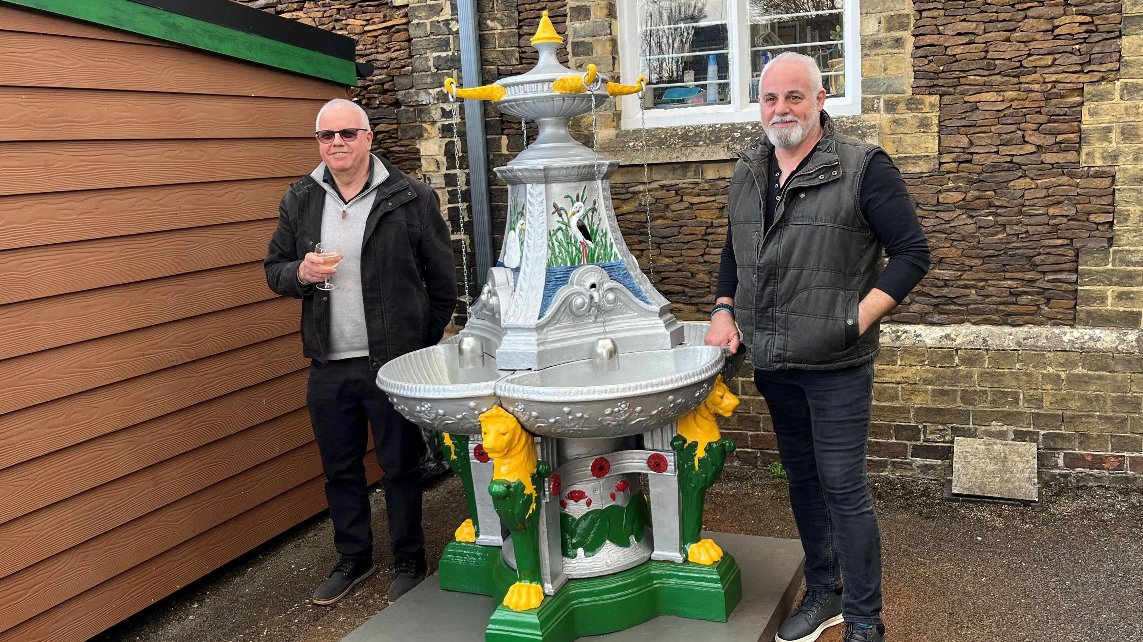 Two men stand either side of a 2m (6ft) high cast iron water feature. Both have grey thinning hair and one has a grey beard and moustache, the other is wearing dark glasses, both dressed casually in jeans with jackets and jumpers. They are in a courtyard of a museum with a wooden shed on the left and an old looking building behind then finished in flint like stone.