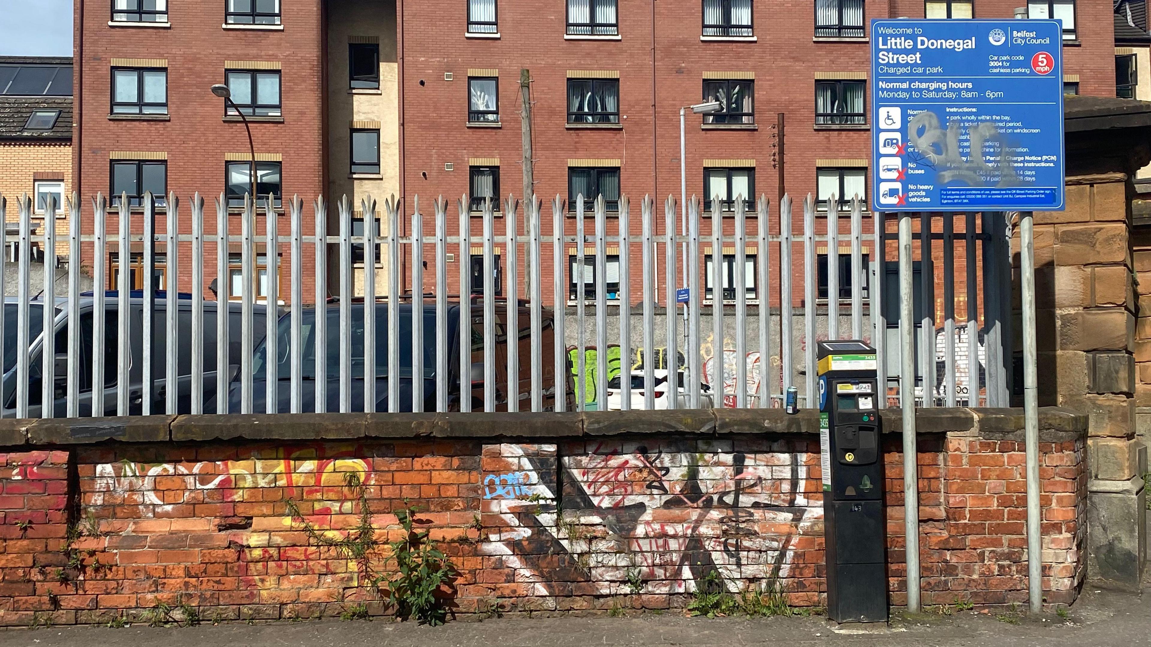 The sign for Little Donegal Street car park. The wall behind is red and is covered in graffiti. There are tall spiked metal gates surrounding the car park.