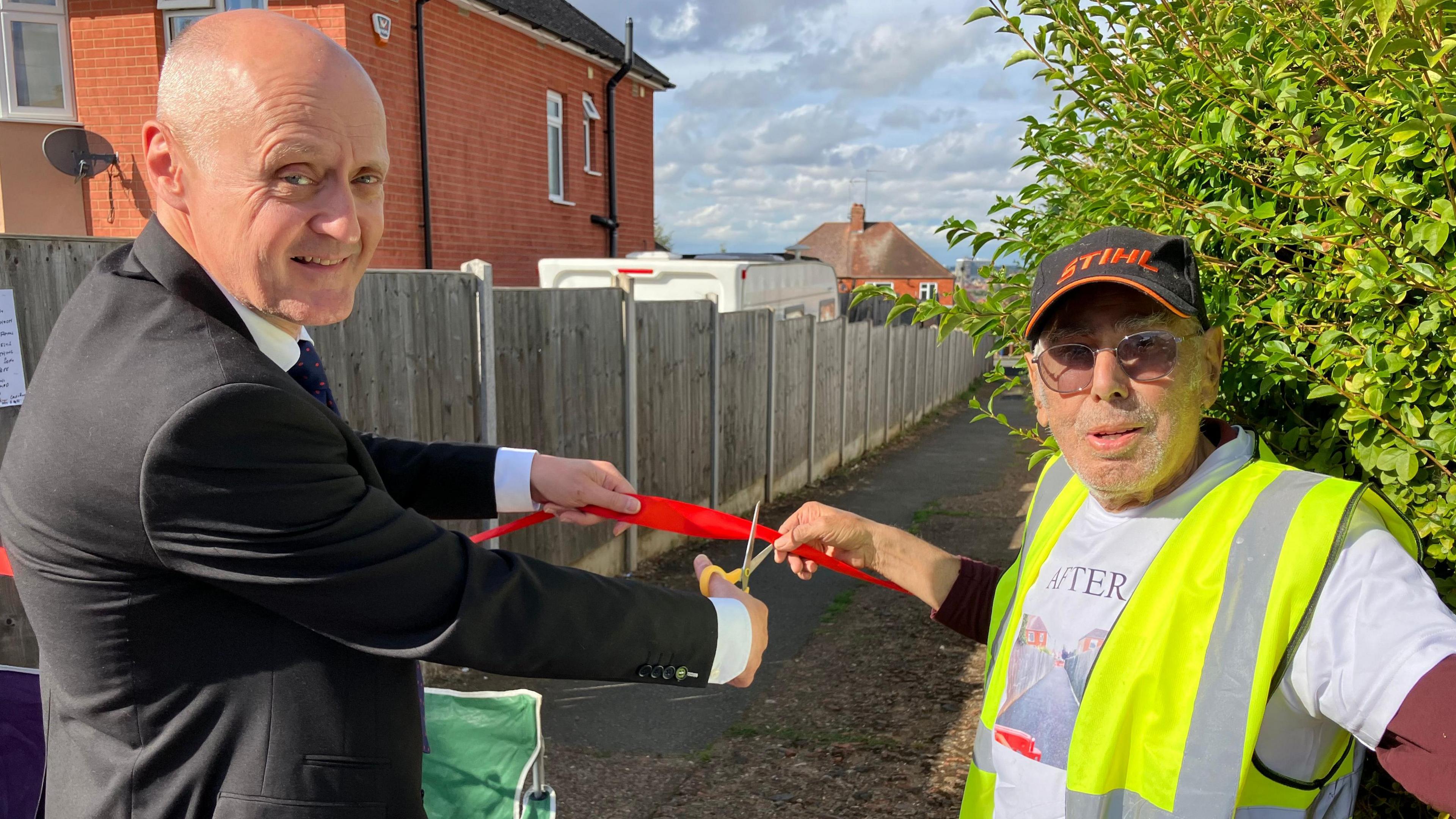 Mike Davey in a black suit about to cut a red ribbon while Chris Antoniou in a white T-shirt holds the ribbon