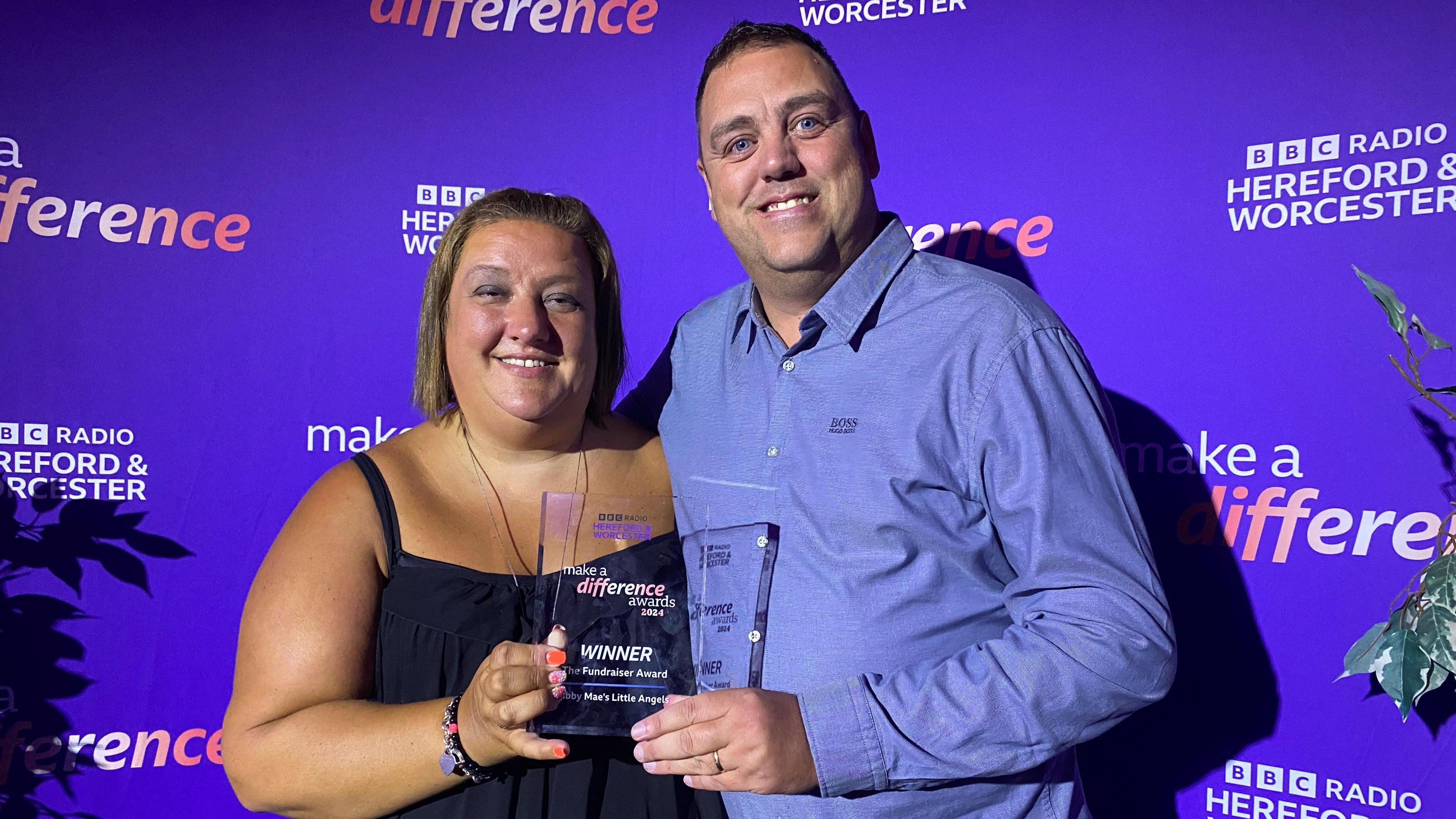 A man and a woman stand in front of a BBC backdrop. They are holding an award. The woman has short brown hair and is wearing a navy top, and the man has short dark hair and is wearing a blue shirt