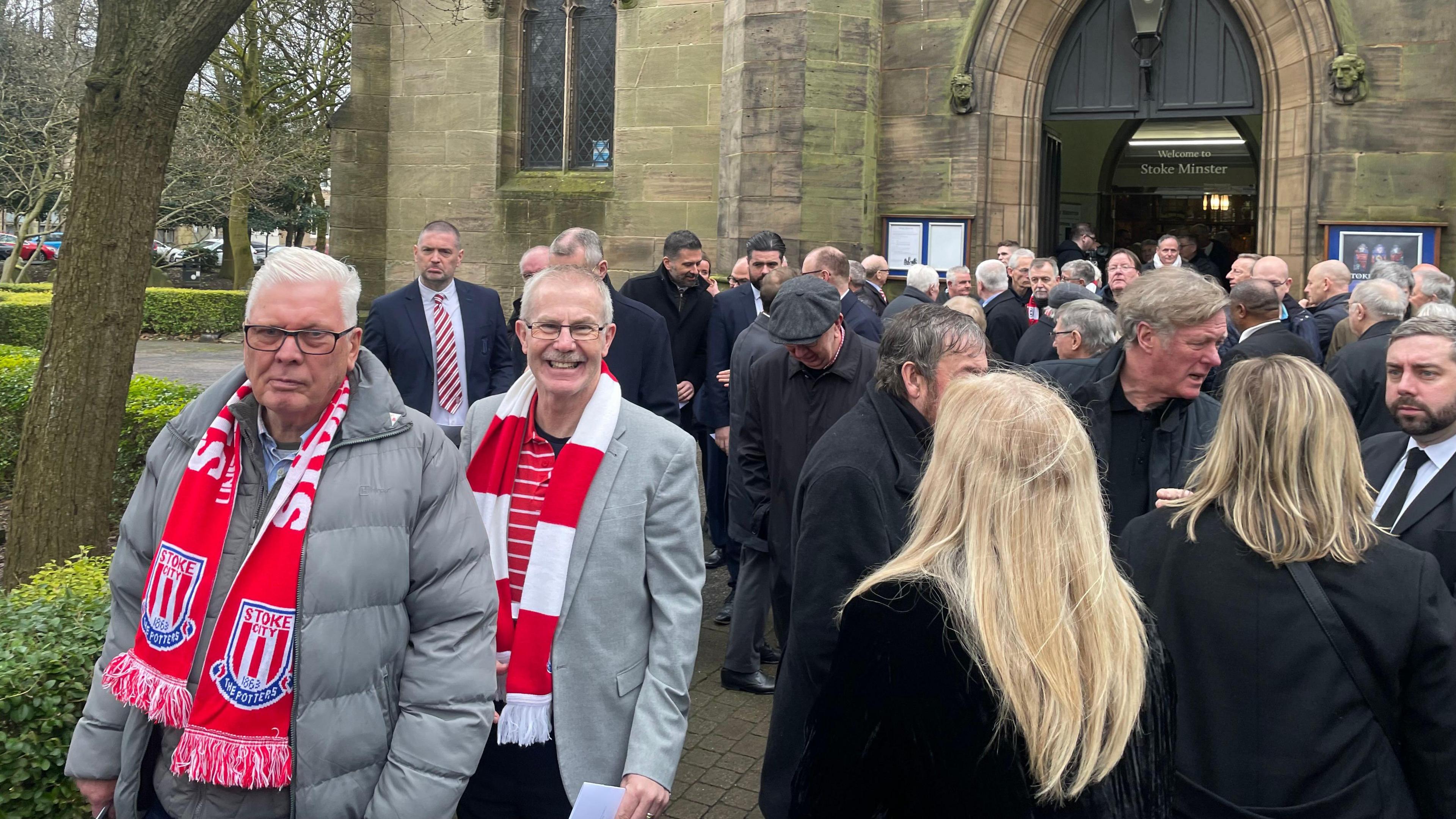 A crowd of people standing in front of a church entrance. Most are wearing dark clothing, including black coats, with two men on the left wearing red and white Stoke City scarves and grey jackets.