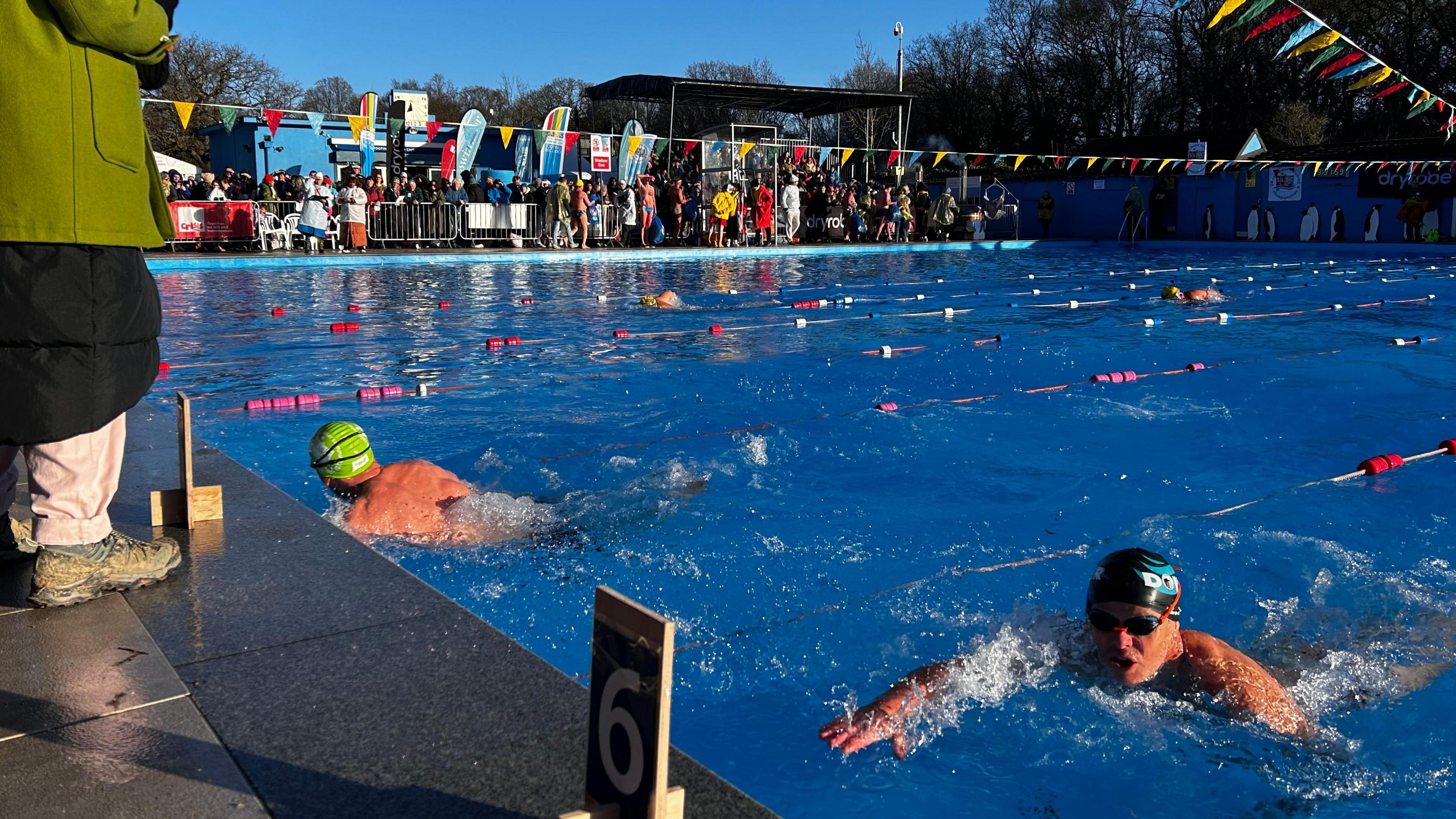 Two swimmers approach the end of the pool at Tooting Bec Lido. A crowd of people watch on.