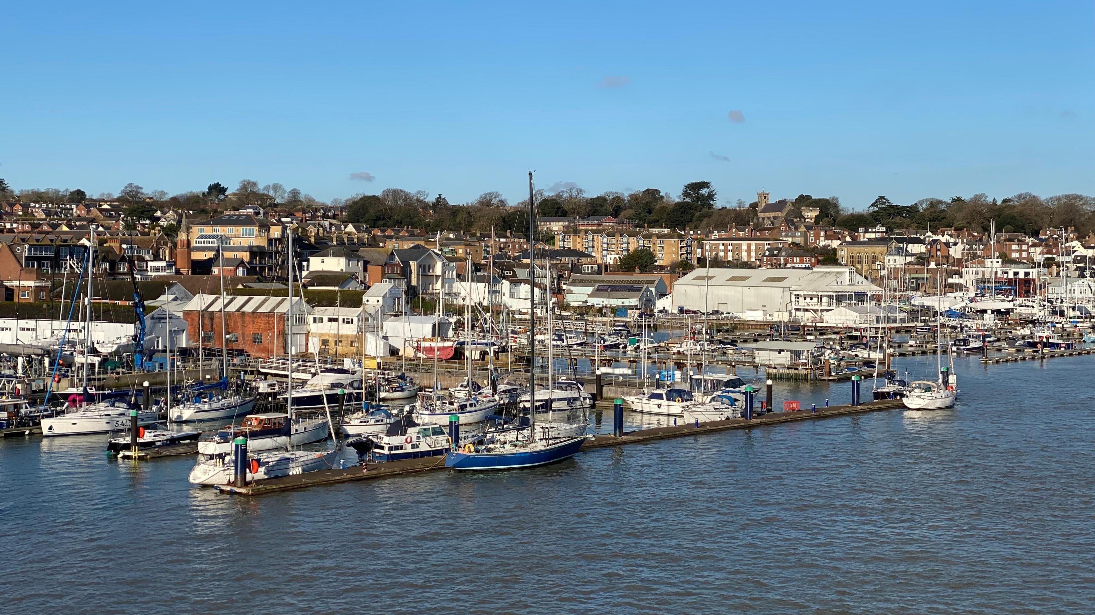 A view over a town from the sea. In the foreground are several moored yachts. Behind you can see industrial buildings and homes. The sky overhead is blue. The photo appears to have been taken from a boat. 