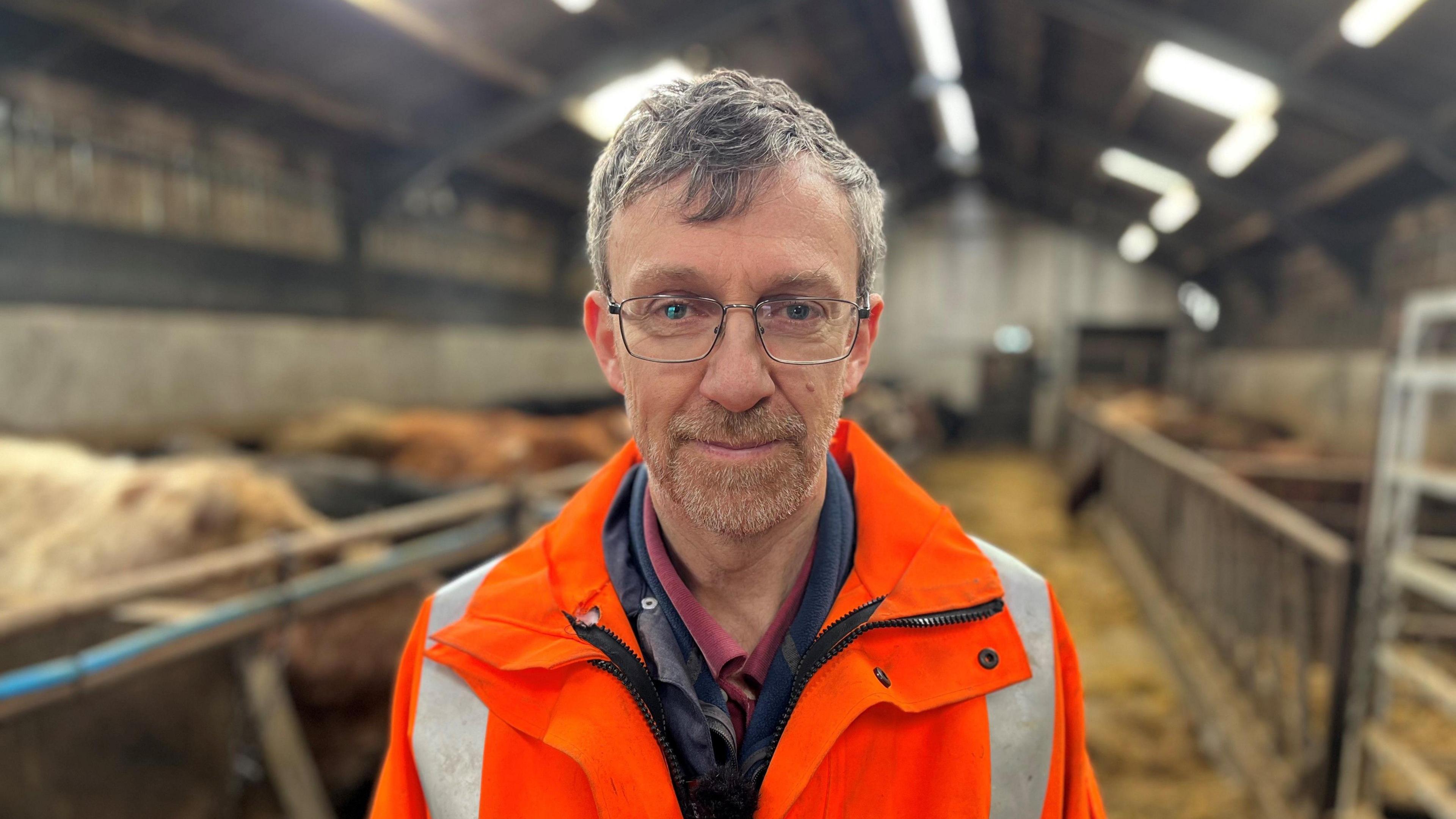 Ian Cursiter, a grey haired bespectacled man wearing a bright orange jacket smiles at the camera in his cow shed