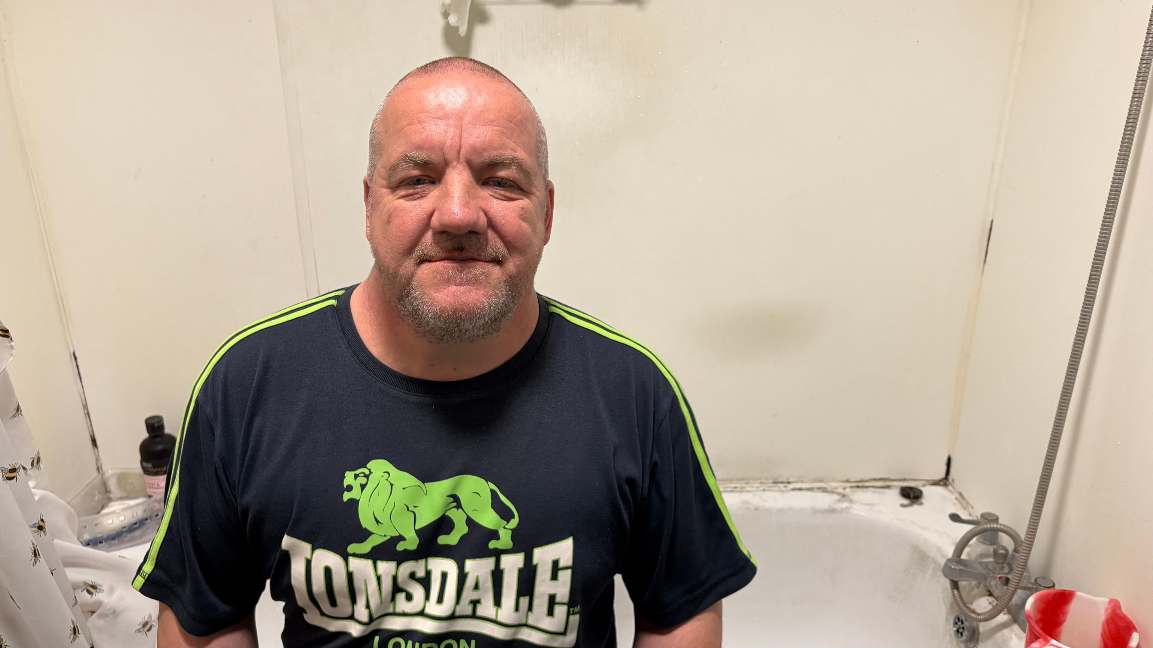 Carl Palmer with very short white hair looking at the camera and wearing a black and green T-shirt with a Lonsdale logo. He is sitting on a bath, and mould can be seen on the wall behind him.
