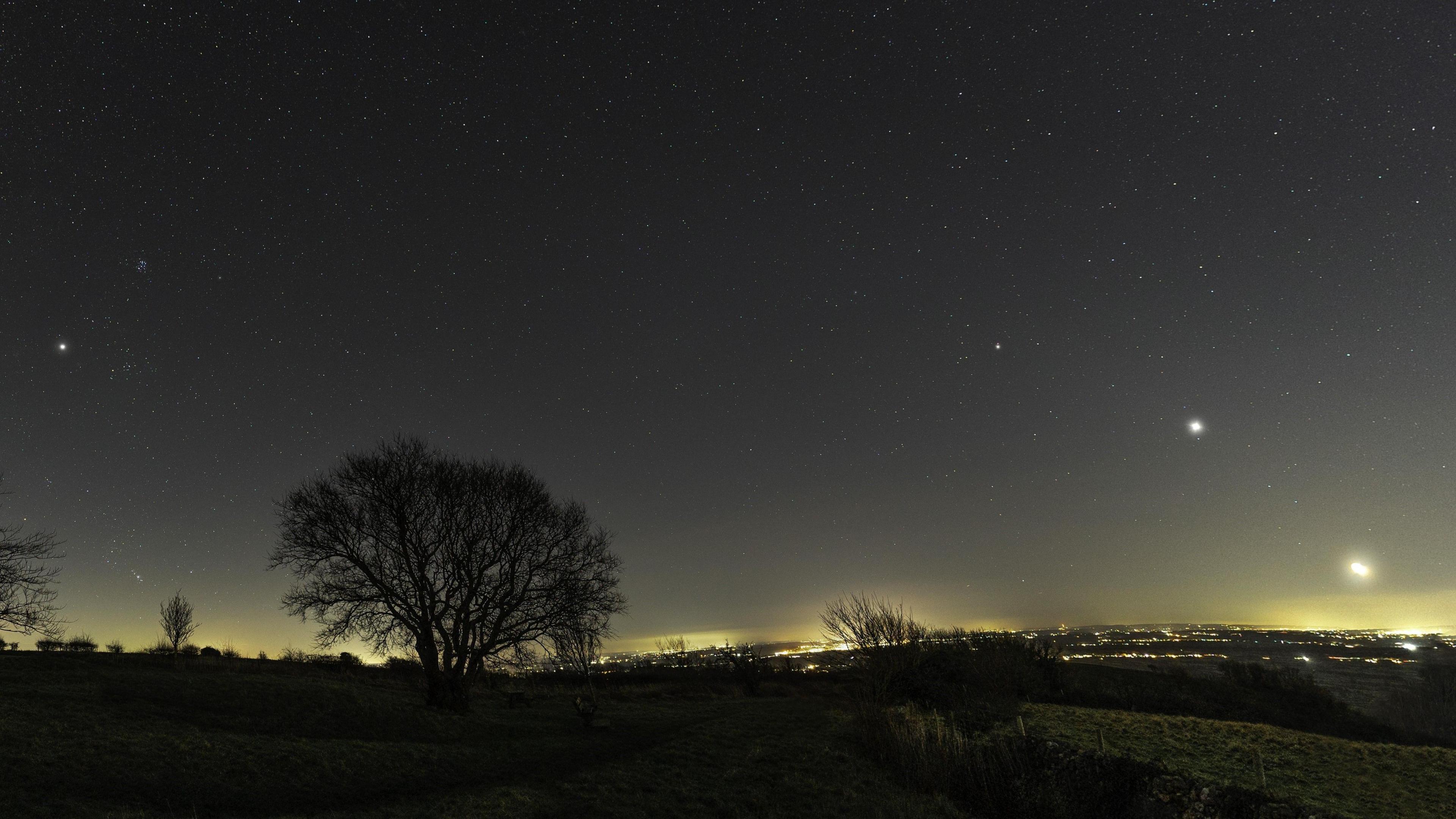 A wide-angle shot of the sky over the Mendip Hills in Somerset taken at night. The sky is dark and various stars are seen brightly shining with trees silhouetted in the foreground and a light haze over Bristol in the distance