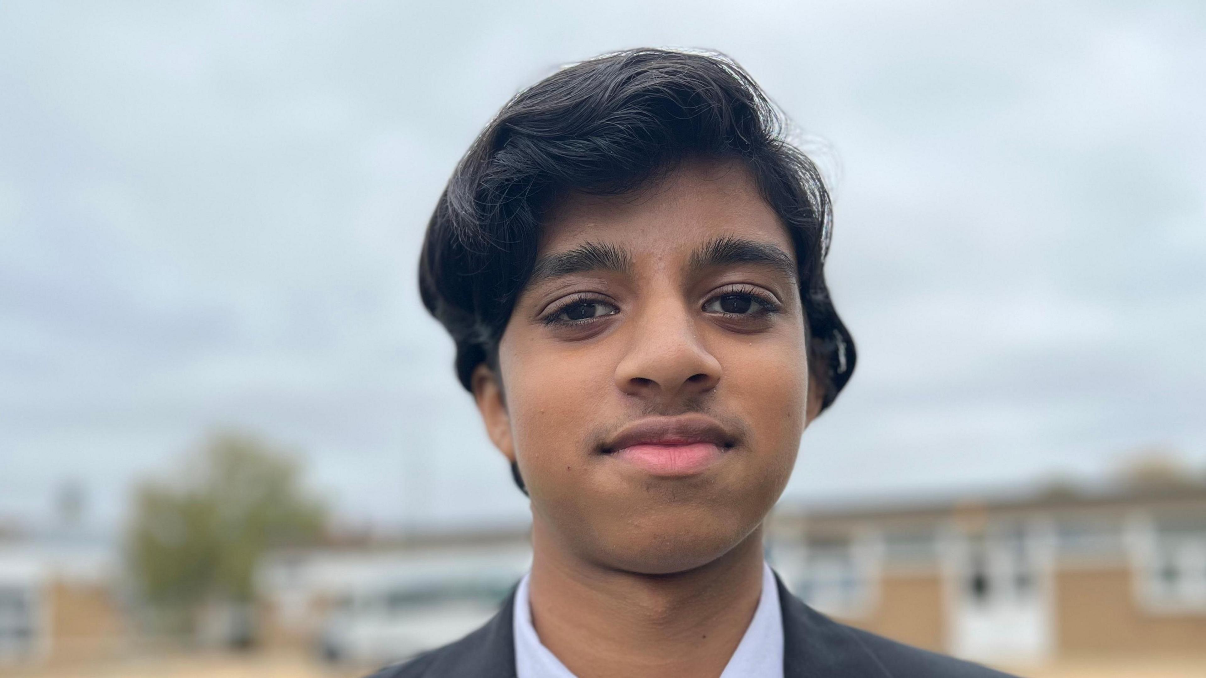 Kamalesh,15 has dark hair, dark eyes and olive skin and is looking at the camera. He is standing in school grounds and wears a black school blazer, white shirt and school tie.