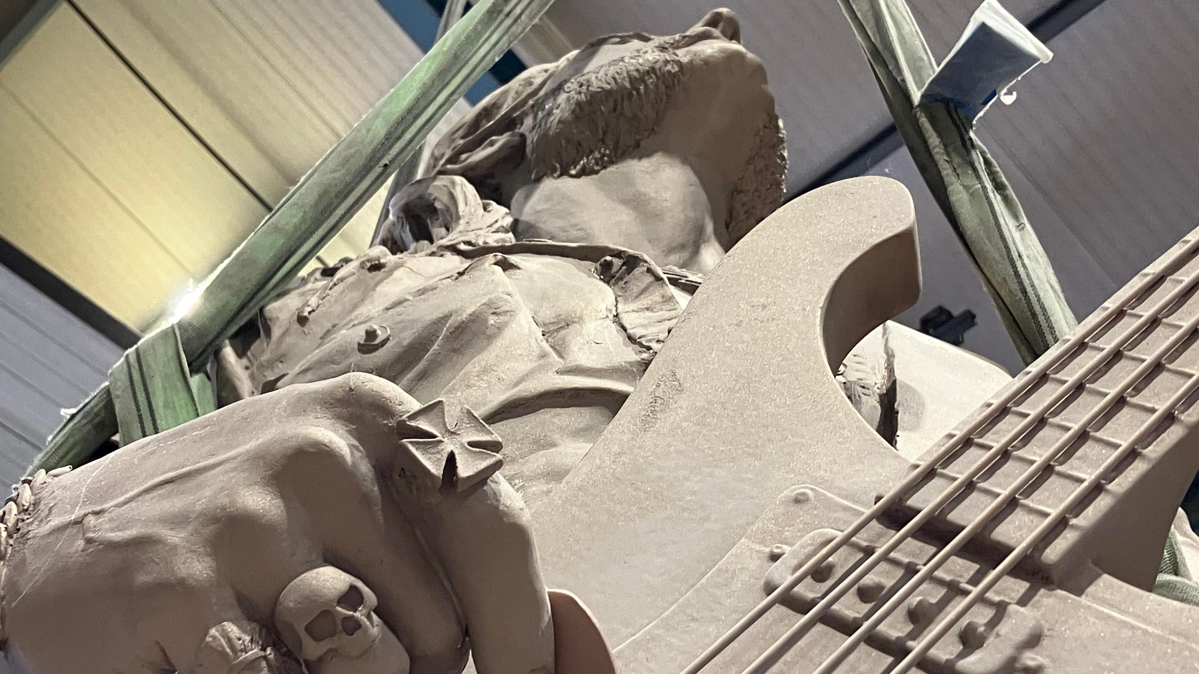 A statue of a man holding a guitar, viewed from below and looking straight up to his chin. The fingers he is using to play the guitar have rings on them, including one of a skull.