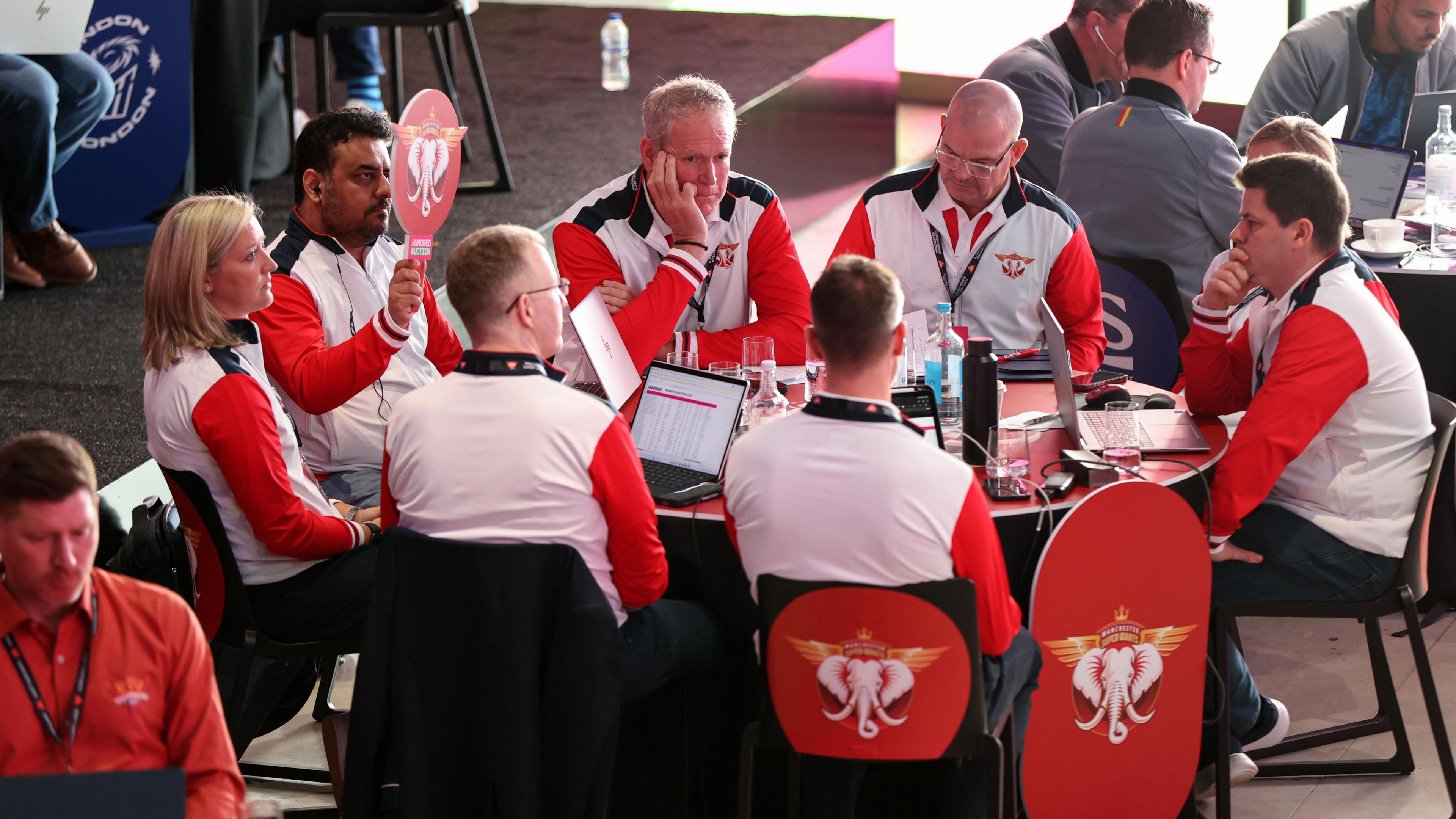The Manchester Super Giants auction team place a bid at the women's 2025 100 auction. They are sat round a table in white and red clothing, and one man holds a padel with an elephant on to signal a bid