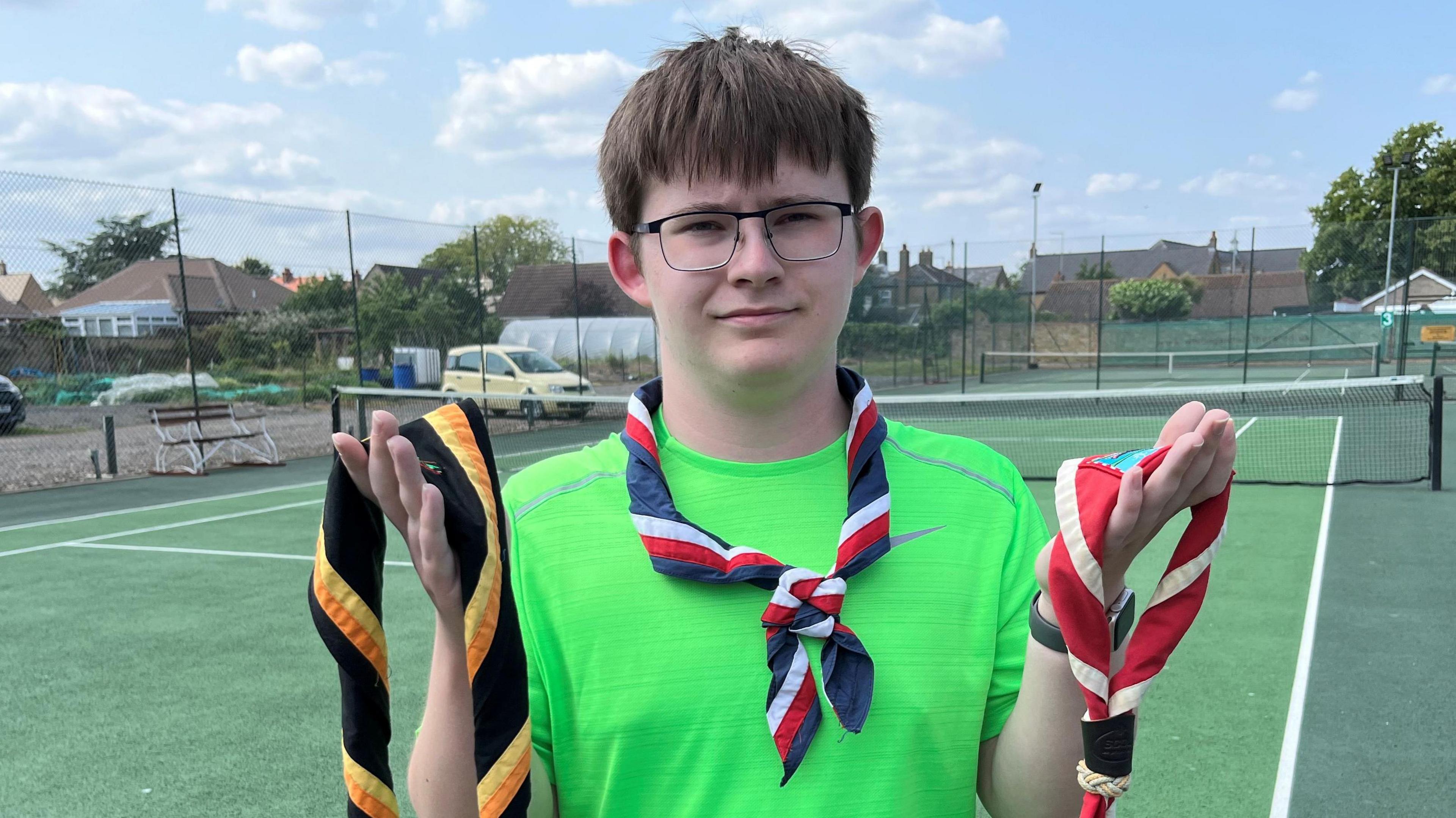Oliver Appleby holding two colourful neckerchiefs and he has one around his neck. Behind him is the hard tennis court.