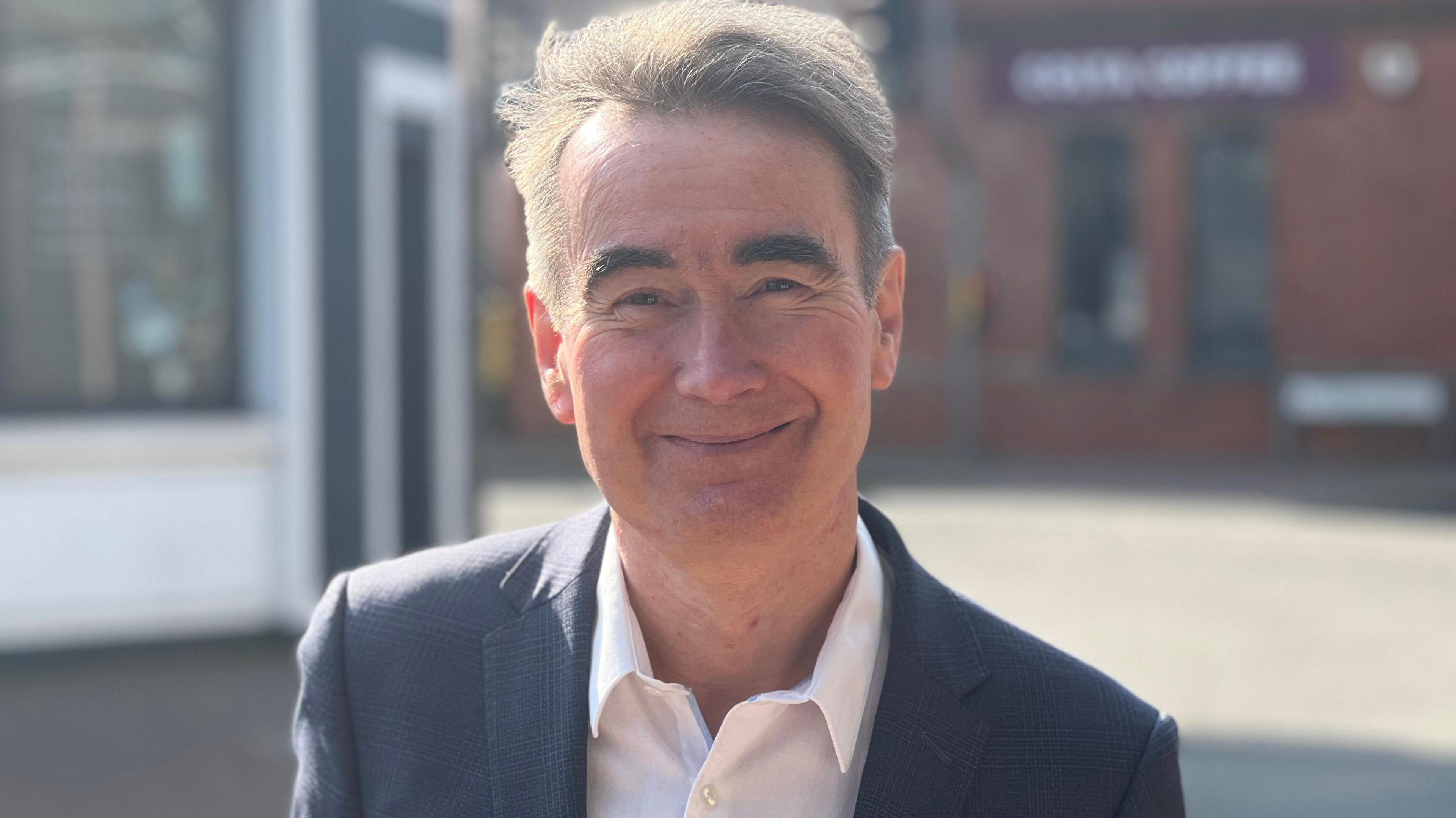 A man with greying hair and wearing a navy jacket with a white shirt, smiling at the camera. He's standing outside with shops in soft focus behind him.