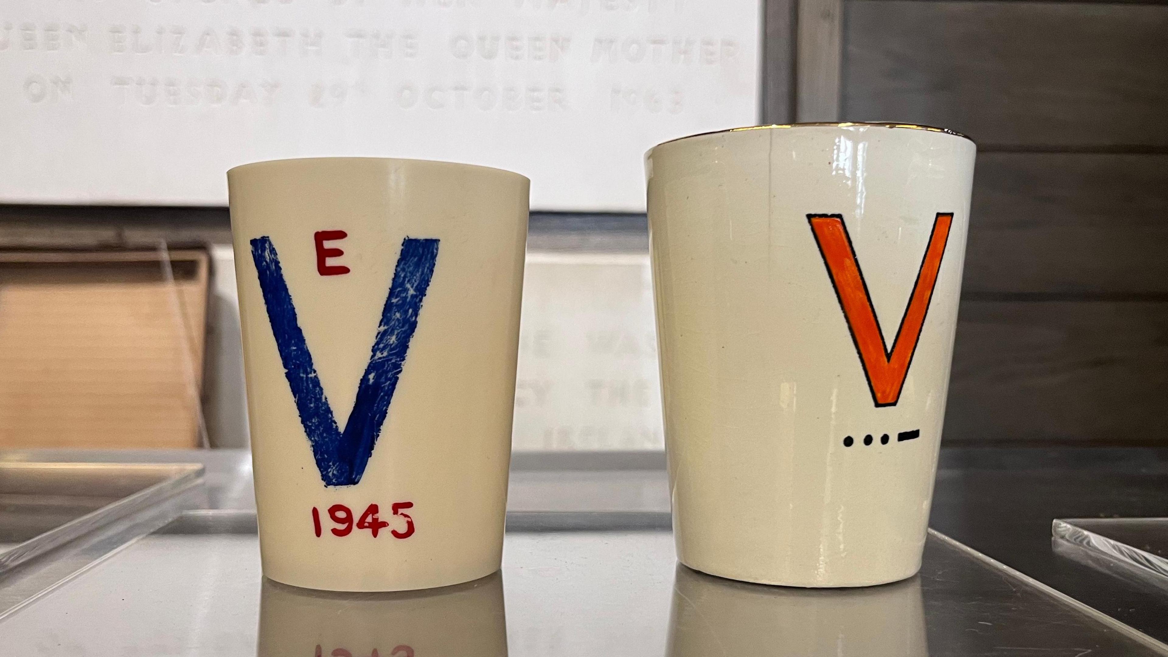 Two cups - both are white. The one on the left is painted with a blue V and the one of the right is painted with a red V.