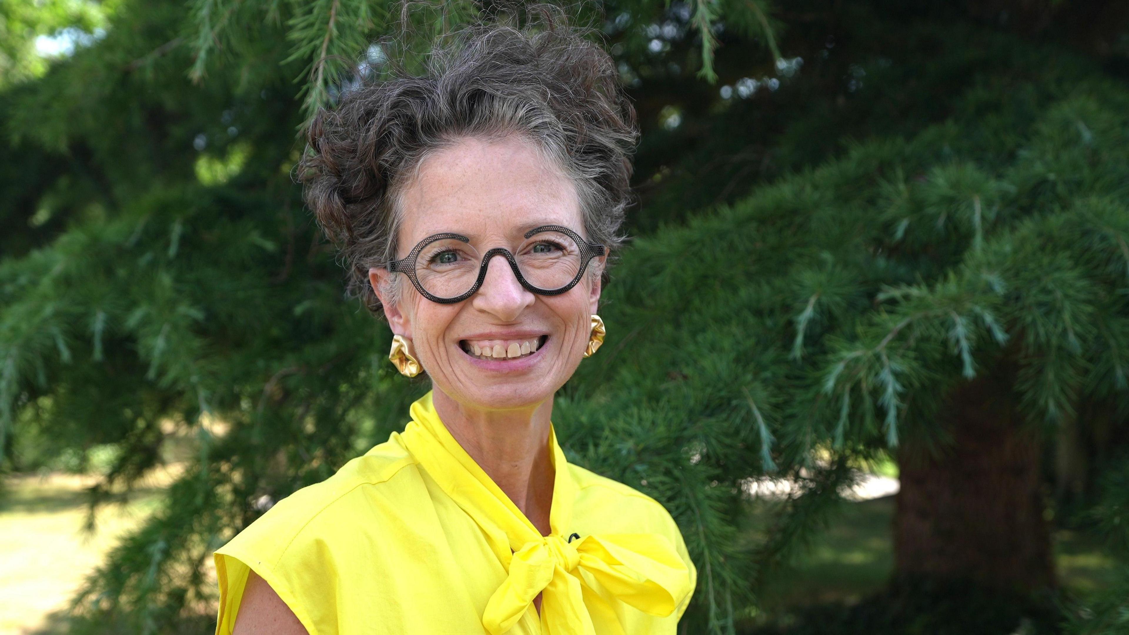 Liz smiles at the camera. She has grey curly short hair and wears glasses with round frames. She wears a bright yellow top and gold statement earrings, and stands in front of a large tree.