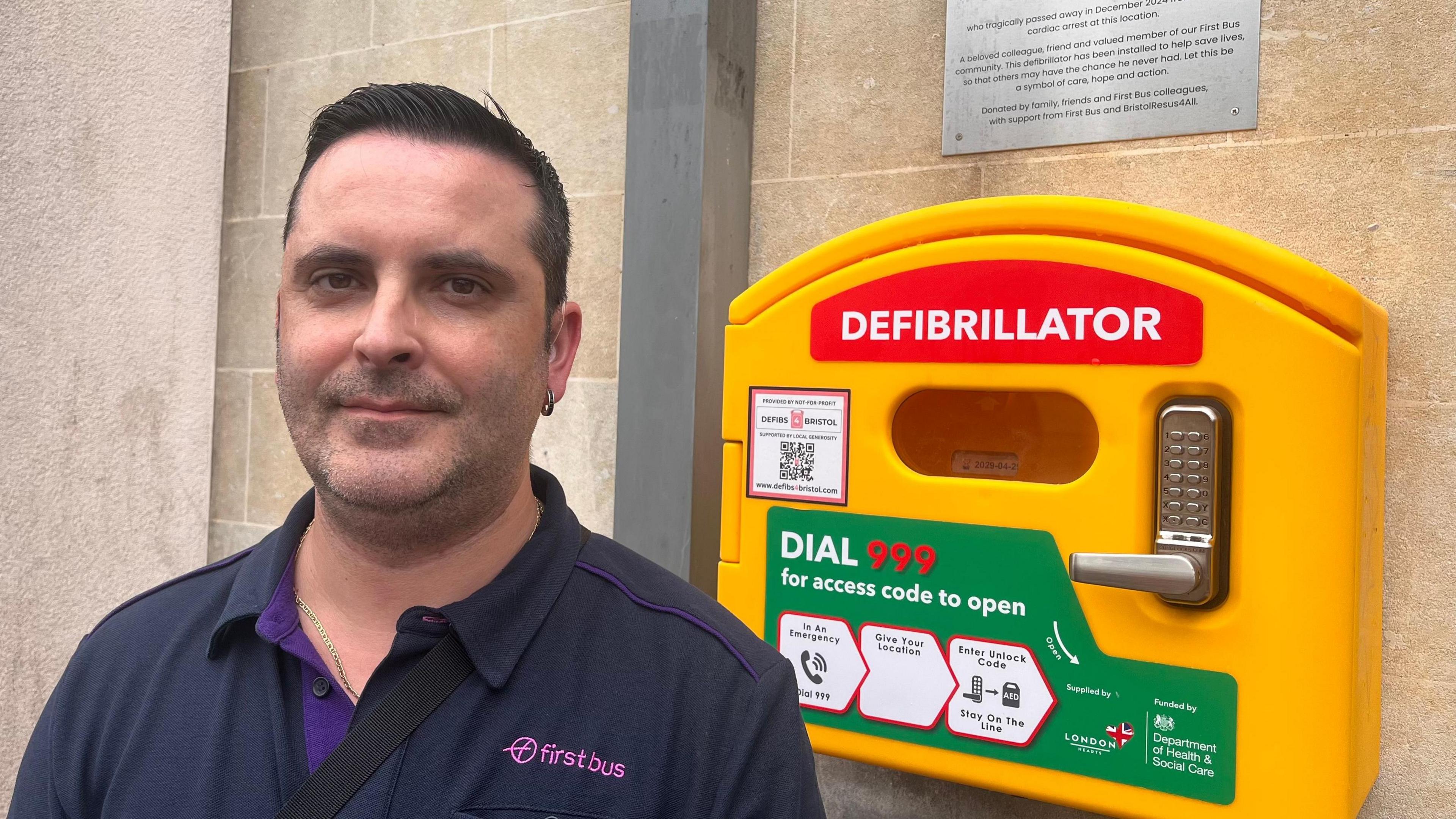 Carlos Santos stands in front of a bright yellow defibrillator cabinet mounted on a brick wall. He is wearing a black shirt. The defibrillator has red signage with the word “Defibrillator” in white, and a green circular “Call 999” instruction label.