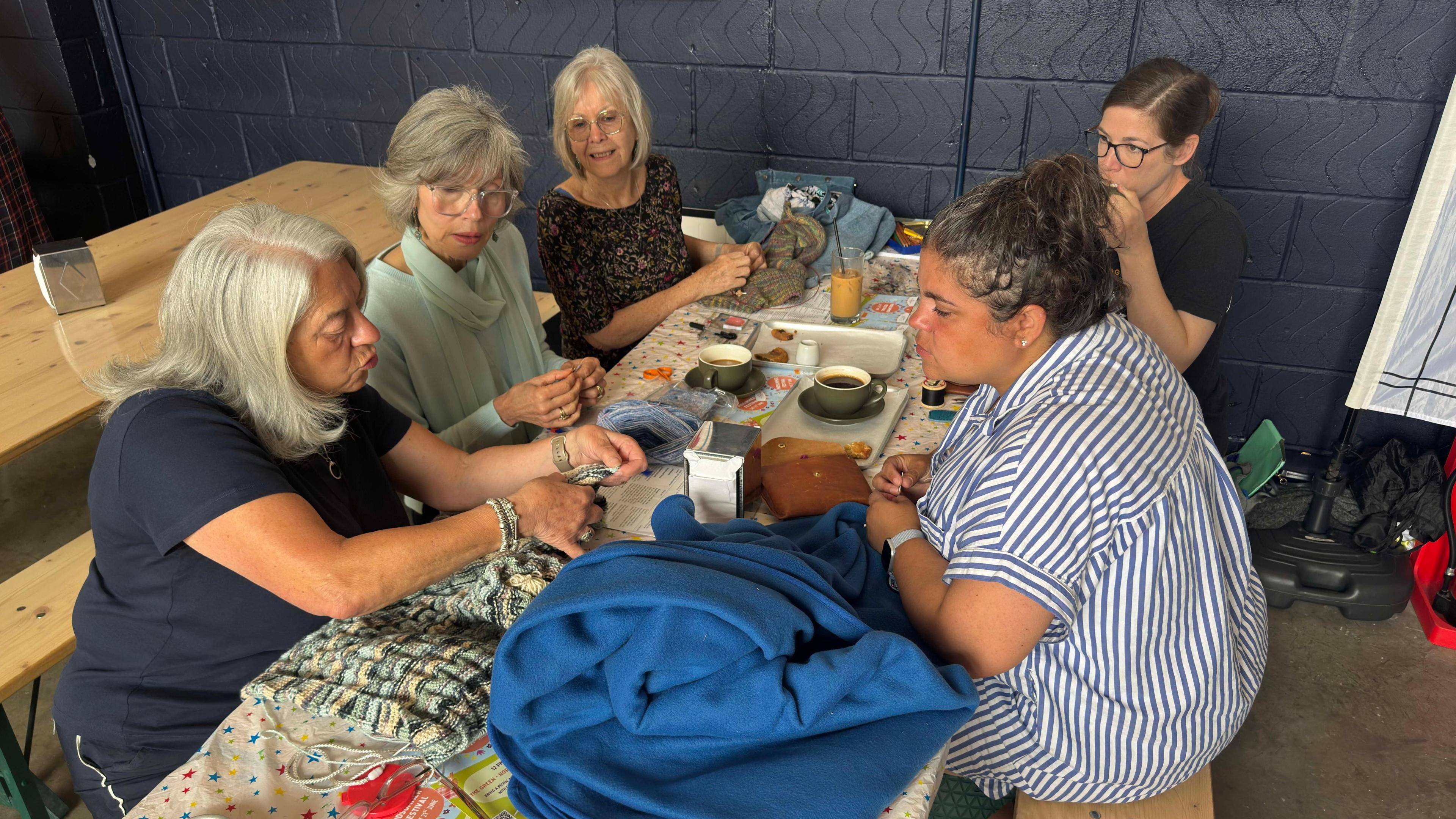 Five women sit around a long table. They are drinking coffee, chatting and doing various crafts, including sewing and knitting. One women is demonstrating a technique to others in the group.