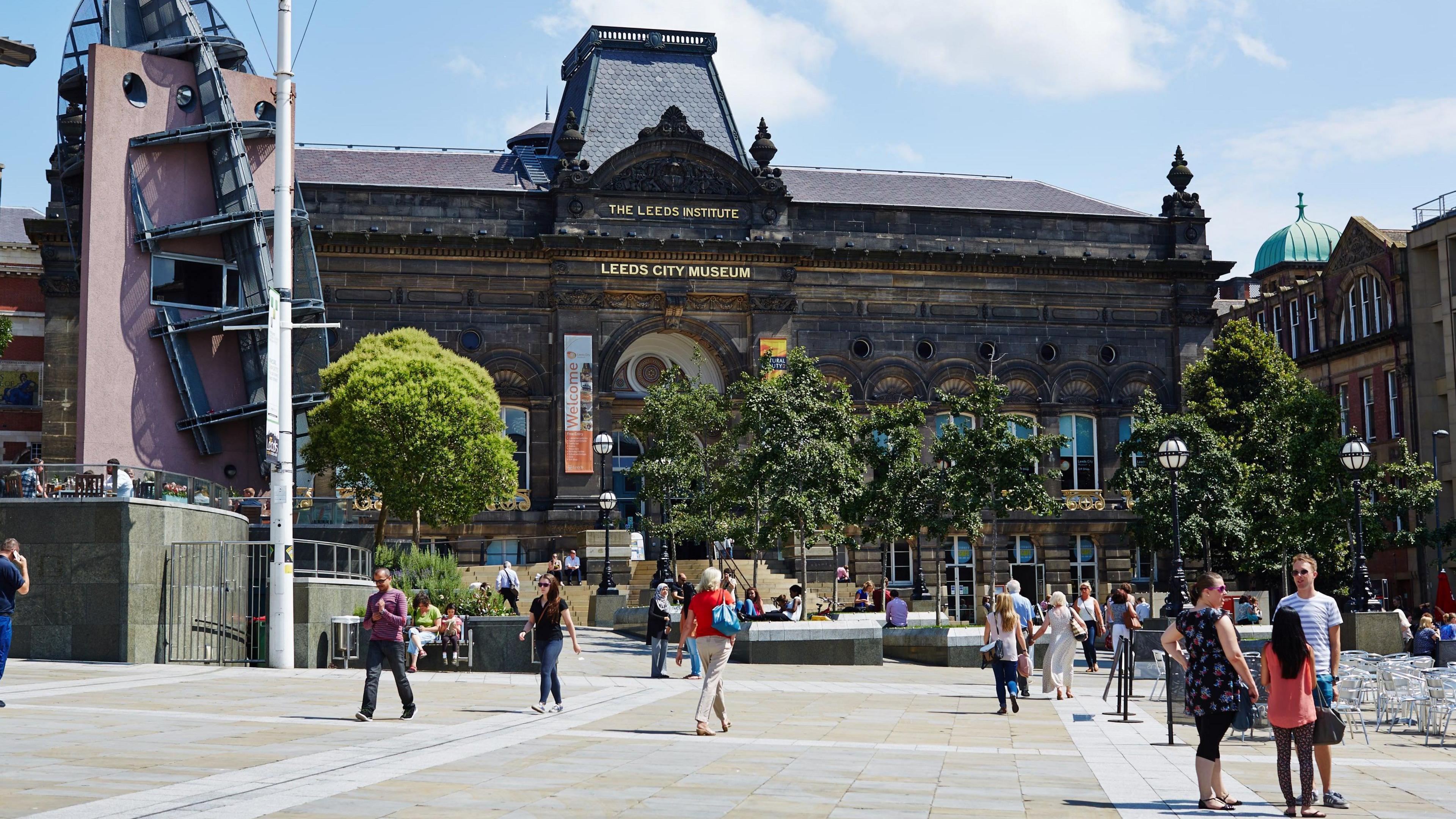 The front of Leeds City Museum. It is a sunny day, with people pictured walking in front of the building.