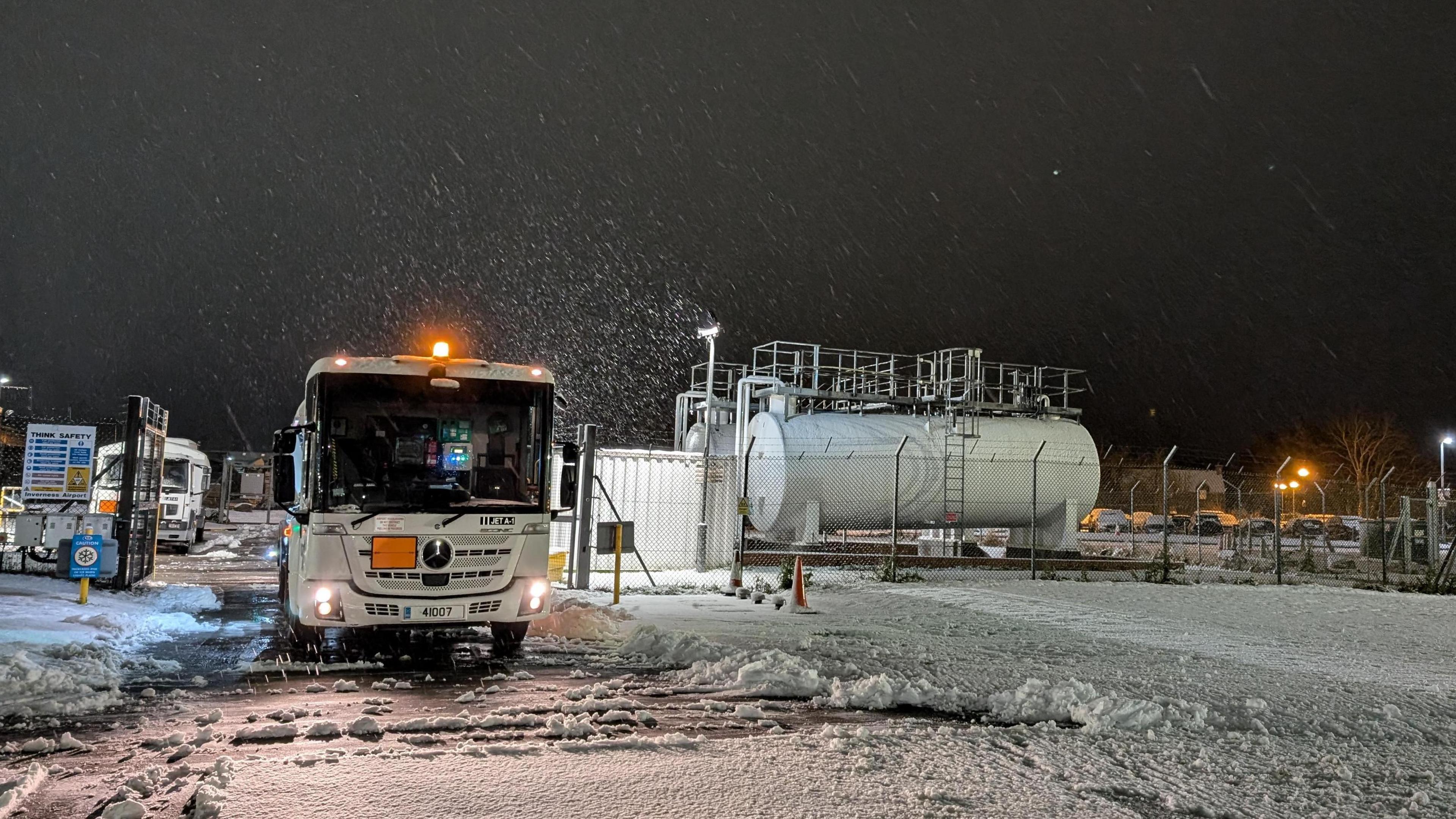 Snow is falling. There is a lorry with an orange flashing light parked near two white fuel tanks. There is another lorry parked in the background.